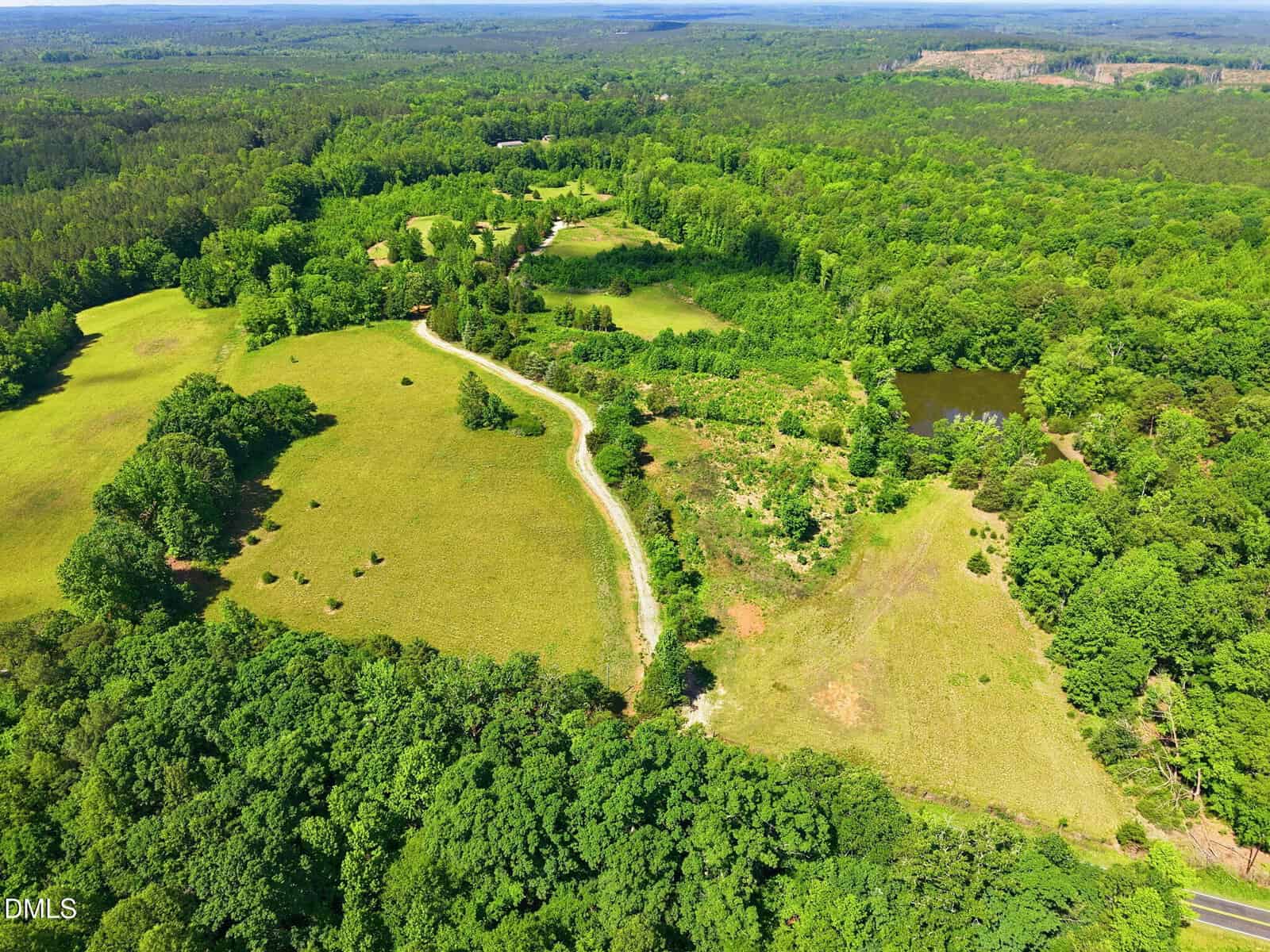 Aerial view of a rural landscape near 001 Keeton Road, featuring open fields, scattered trees, a winding dirt road, and a small pond surrounded by dense green forest under a clear sky in Bullock.