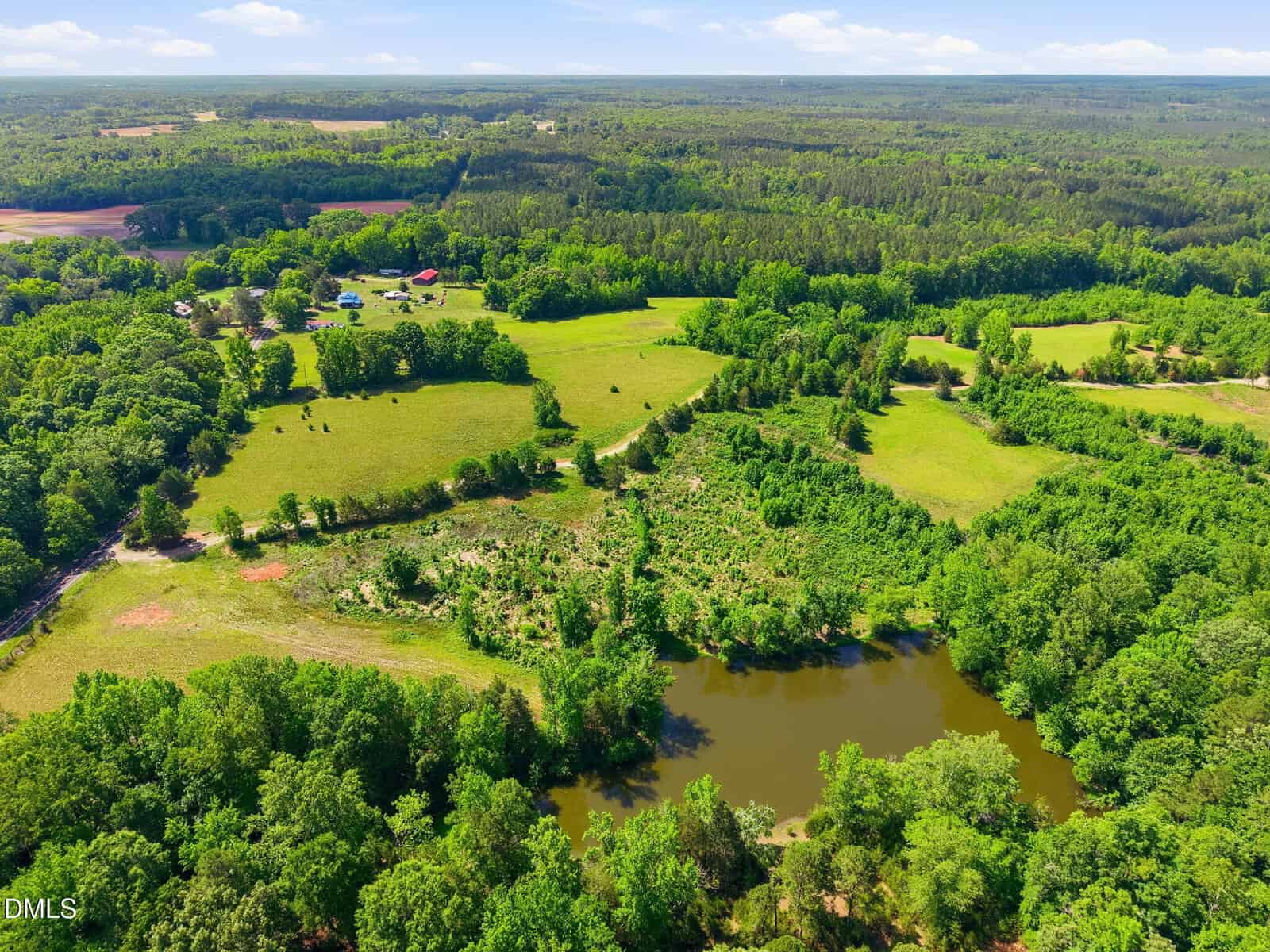 Aerial view of a rural landscape featuring green fields, patches of trees, a pond, and several farm buildings in the distance under a blue sky with scattered clouds.
