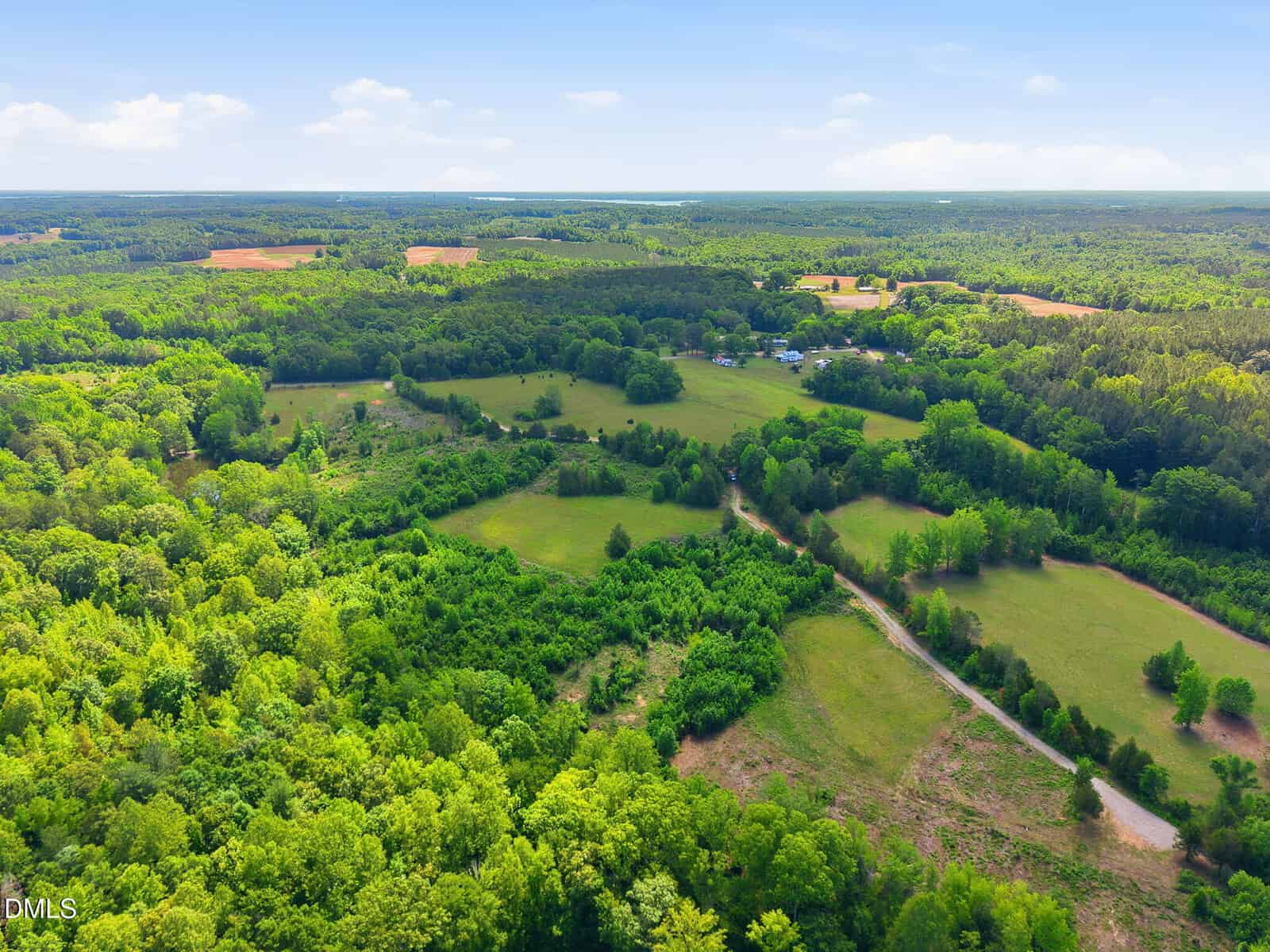 Aerial view of a lush, green rural landscape with rolling hills, patches of forest, open fields, and a few scattered buildings under a blue sky with scattered clouds.