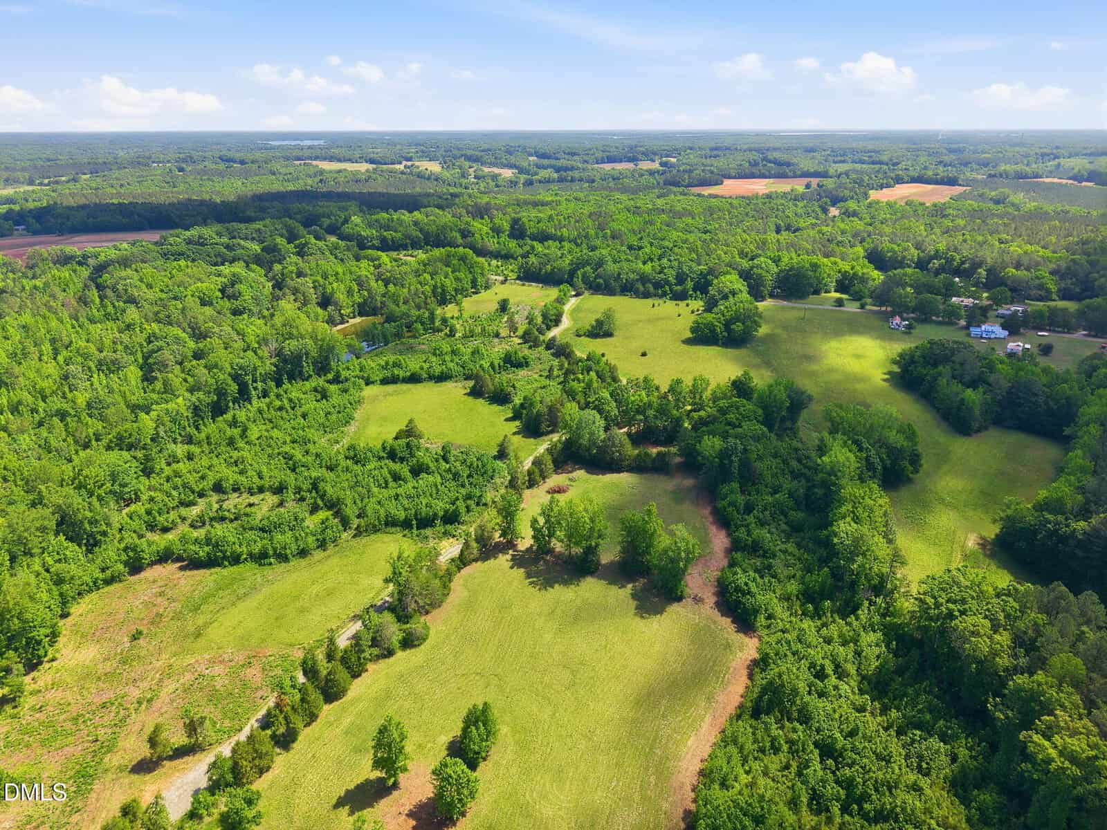 Aerial view of a lush, green rural landscape with rolling hills, open fields, clusters of trees, and a few scattered homes under a partly cloudy sky.