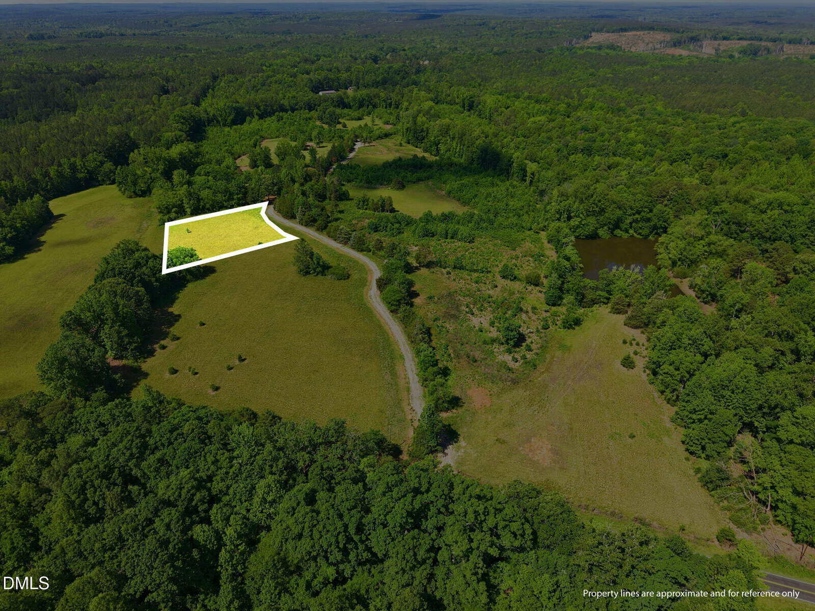 Aerial view of a green, wooded landscape with a winding dirt road and an outlined rectangular plot of land in a field, surrounded by trees and near a pond. The plot is highlighted in yellow.