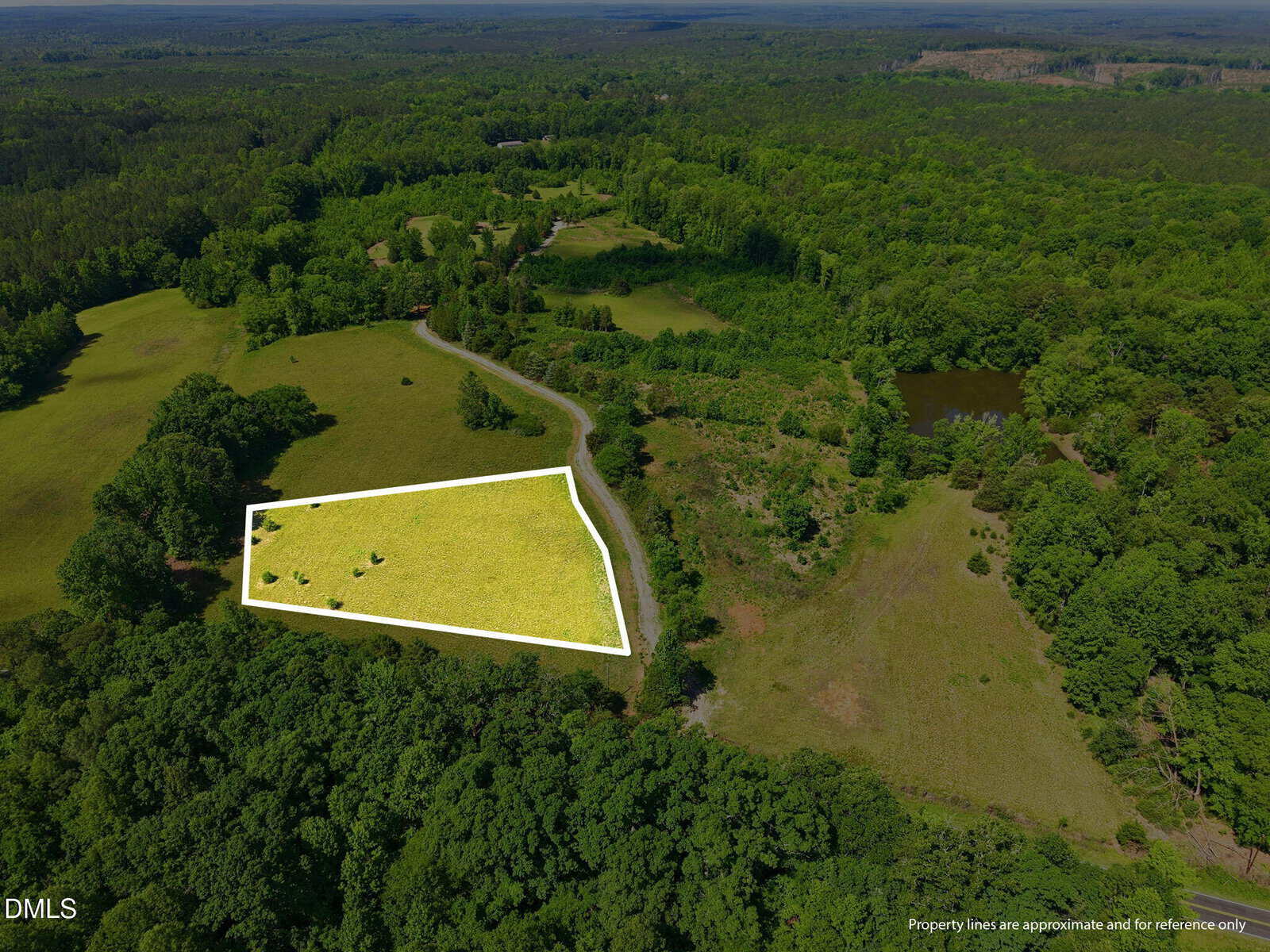 Aerial view of a rural landscape with green fields, trees, and a pond. A section of land is highlighted with a white outline, indicating approximate property boundaries. A road curves along one edge of the marked area.