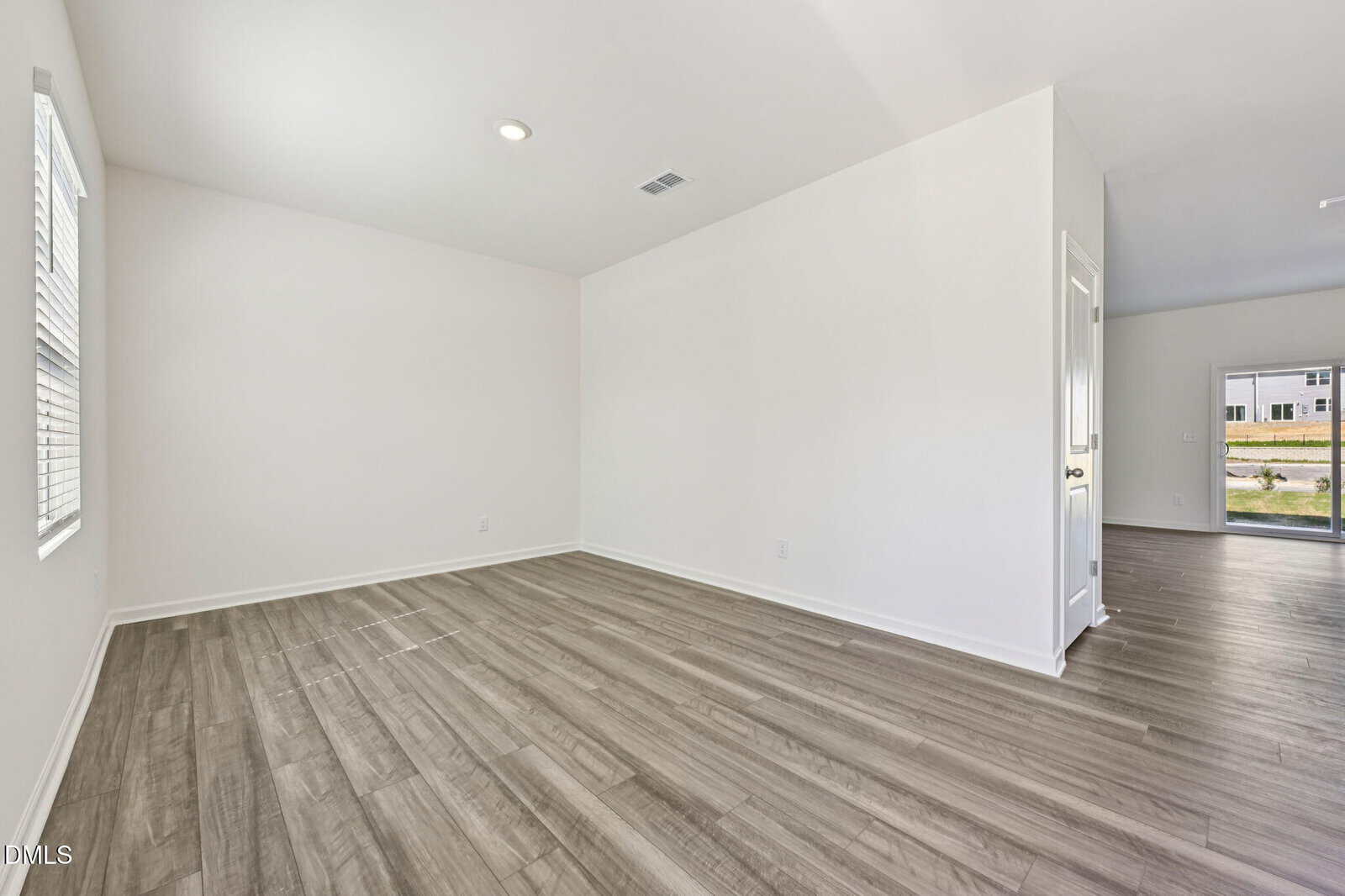 A bright, empty room with light-colored walls, wood-style flooring, two windows, and a recessed ceiling light. An open doorway leads to another area with a view of the outdoors.