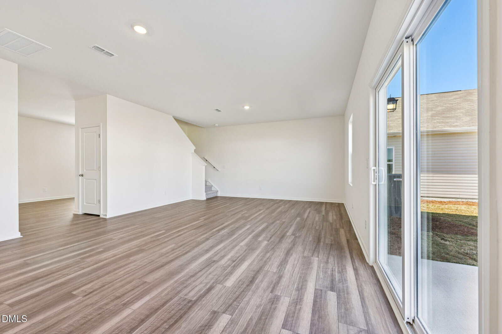 Spacious empty living room with light wood flooring, white walls, large window, sliding glass door to the outside, and stairs leading to an upper floor.