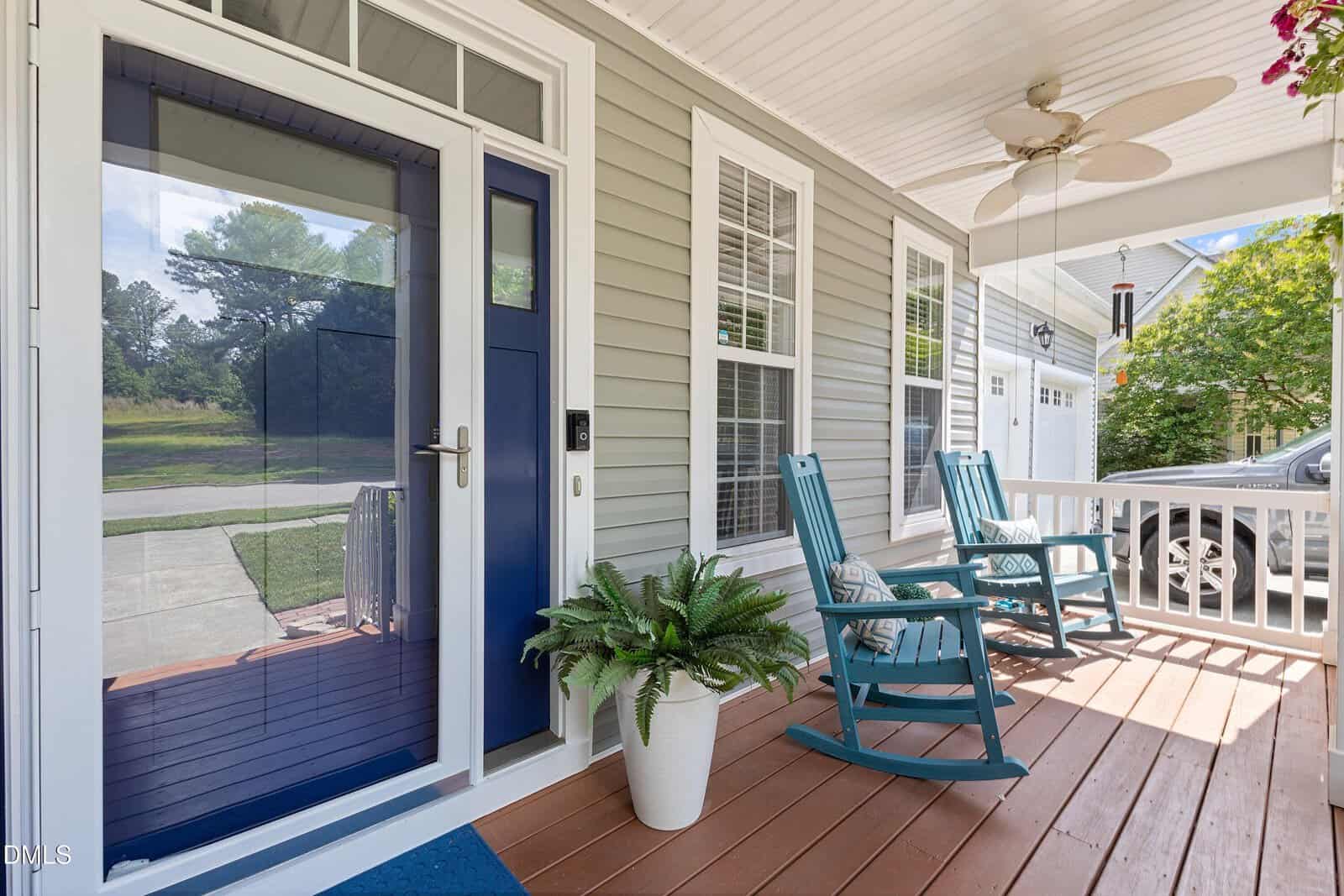 A cozy front porch with two blue rocking chairs, a ceiling fan, a potted fern, and white railings. The porch has brown wooden flooring and overlooks a green yard and quiet street.