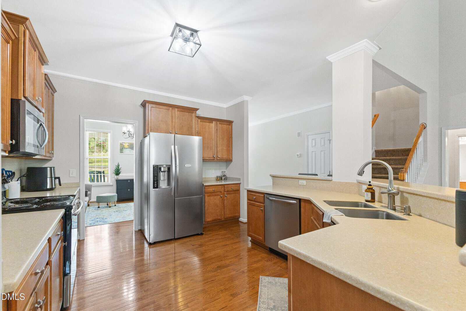 A bright, spacious kitchen with wood cabinets, stainless steel appliances, light countertops, and hardwood floors. An open doorway leads to a sunlit living area, and a staircase is visible in the background.
