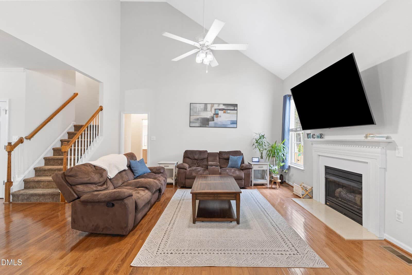 Bright living room with two brown sofas, a wooden coffee table on a patterned rug, a fireplace with a large TV above it, houseplant by the window, and stairs leading to an upper floor. High vaulted ceiling with a ceiling fan.