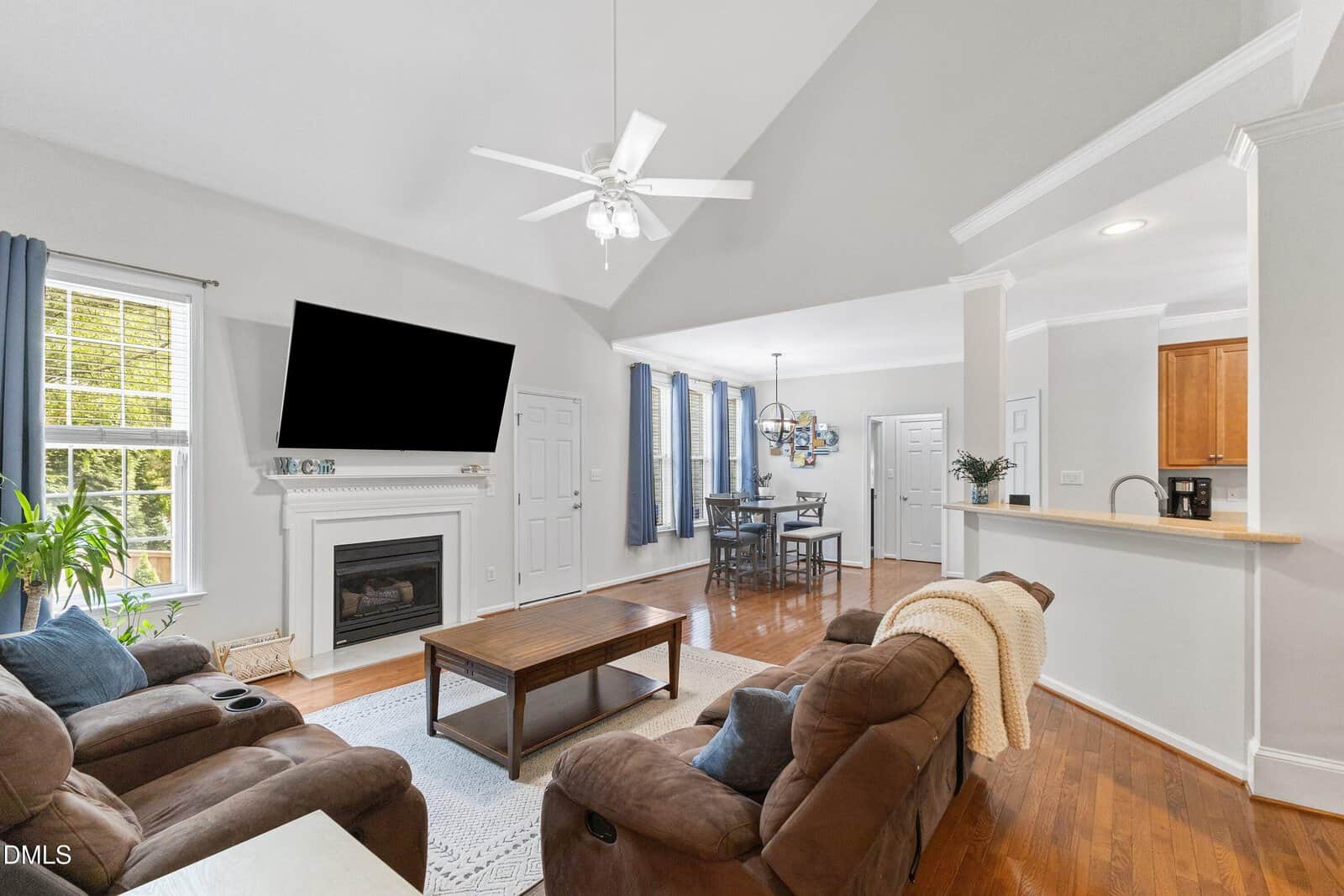 A bright living room with vaulted ceiling, brown sofas, a coffee table, a fireplace with a TV above it, hardwood floors, and large windows. The space opens to a dining area and a kitchen with a breakfast bar.