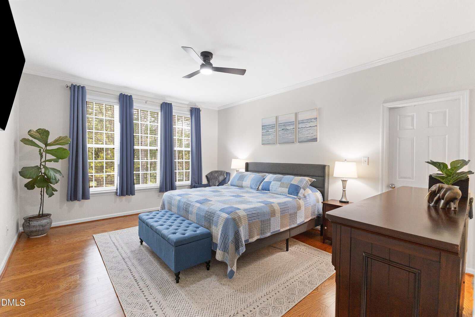 Bright bedroom with large windows, blue curtains, a bed with a blue plaid comforter, blue tufted bench, wooden dresser, white walls, hardwood floors, potted plants, and ceiling fan.