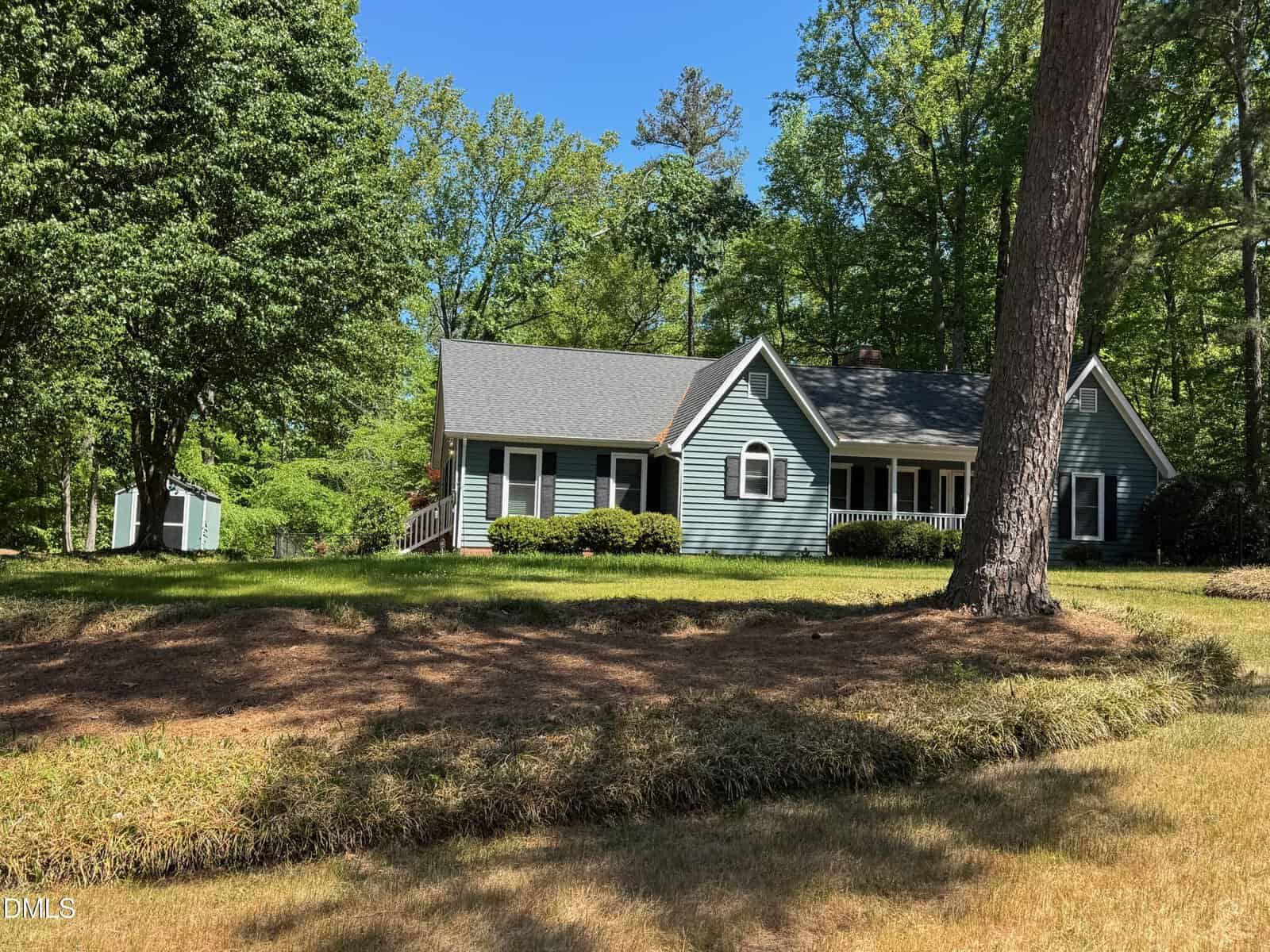 A blue-gray single-story house with white trim and a covered porch sits among tall trees and green grass at 161 Shadow Brook Drive in Henderson. Shrubs line the front, and a small shed is visible in the background on this sunny day.