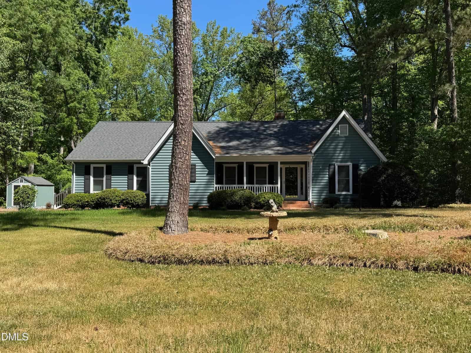 Single-story blue house with white trim and a covered front porch, surrounded by trees and greenery. A small birdbath or garden ornament is in the front yard, and a shed is visible on the left side of the image.