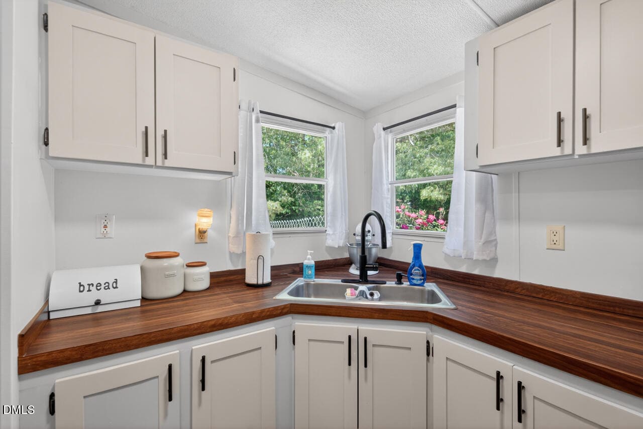 A bright kitchen corner with two windows above a sink, brown wooden countertops, white cabinets, and items like a bread box, jars, and soap on the counter. Trees and flowers are visible outside.