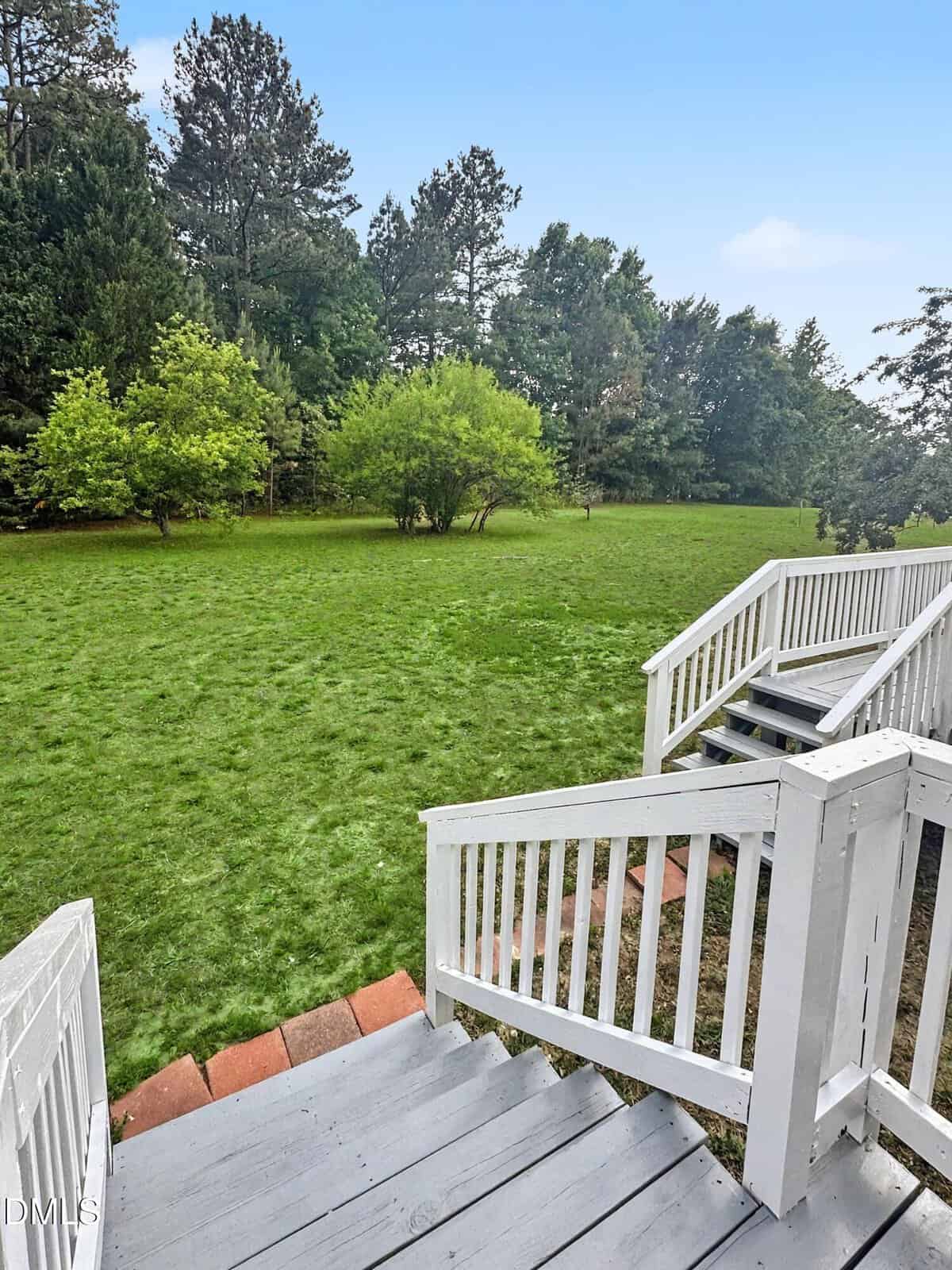 View from a white wooden deck staircase leading down to a spacious, grassy backyard bordered by trees and bushes under a clear blue sky.