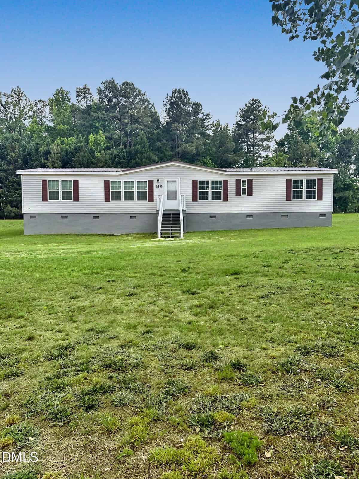 A single-story manufactured home with gray siding, maroon shutters, and a small front porch with white steps, set on a large grassy lawn with a backdrop of trees.