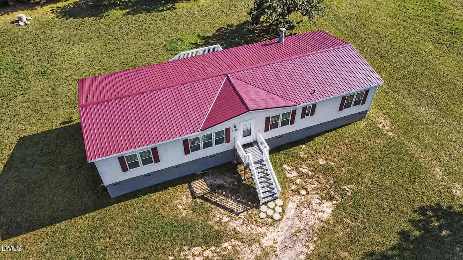 Aerial view of a white, single-story house with a red metal roof, central entrance with stairs, and a large grassy yard surrounding the home.