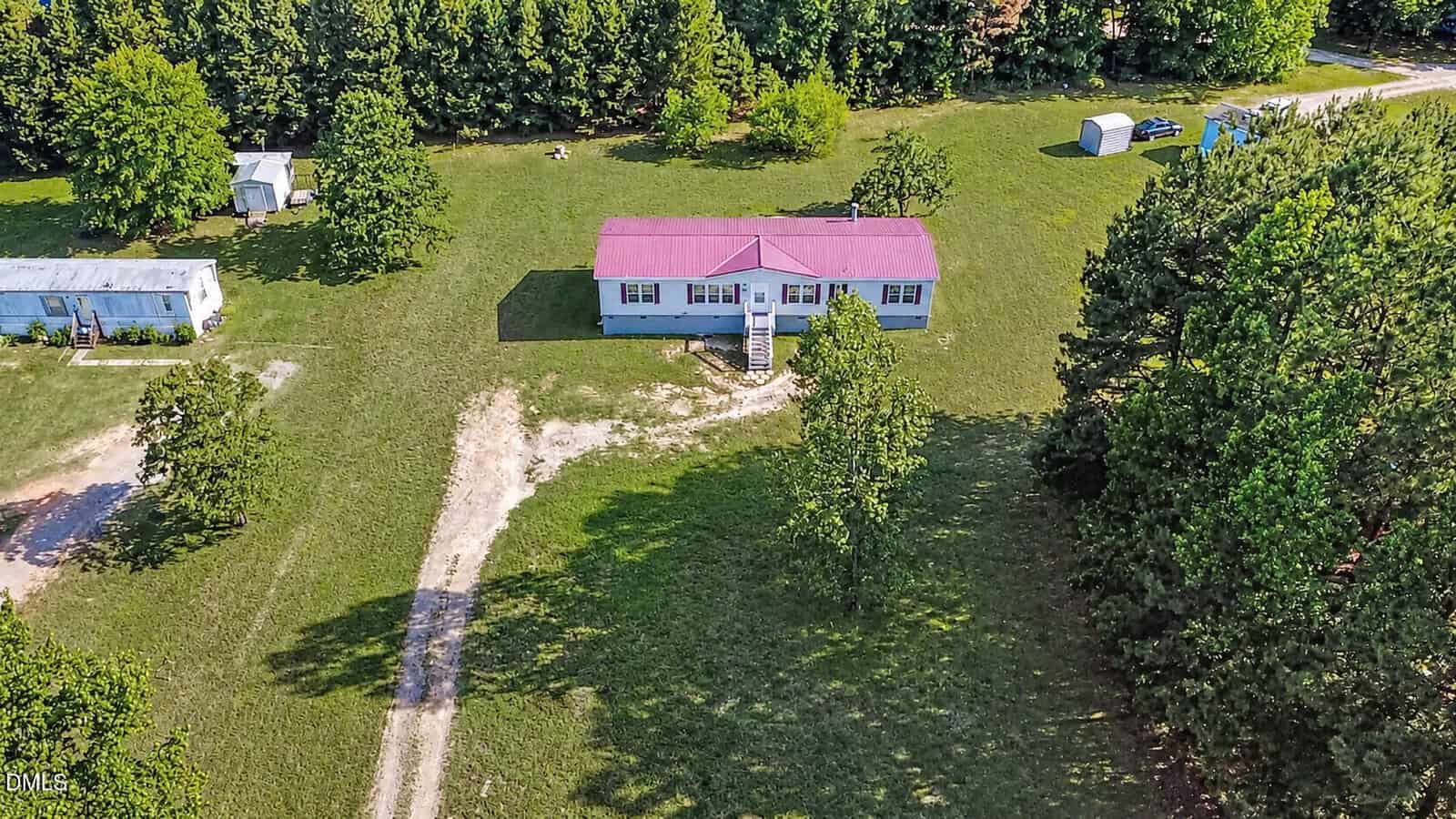 Aerial view of a single-story house with a red roof at 150 Freedom Lane, Henderson. Front steps lead to a large grassy yard bordered by trees, and a gravel driveway curves from the lower edge toward the house, with other small structures nearby.