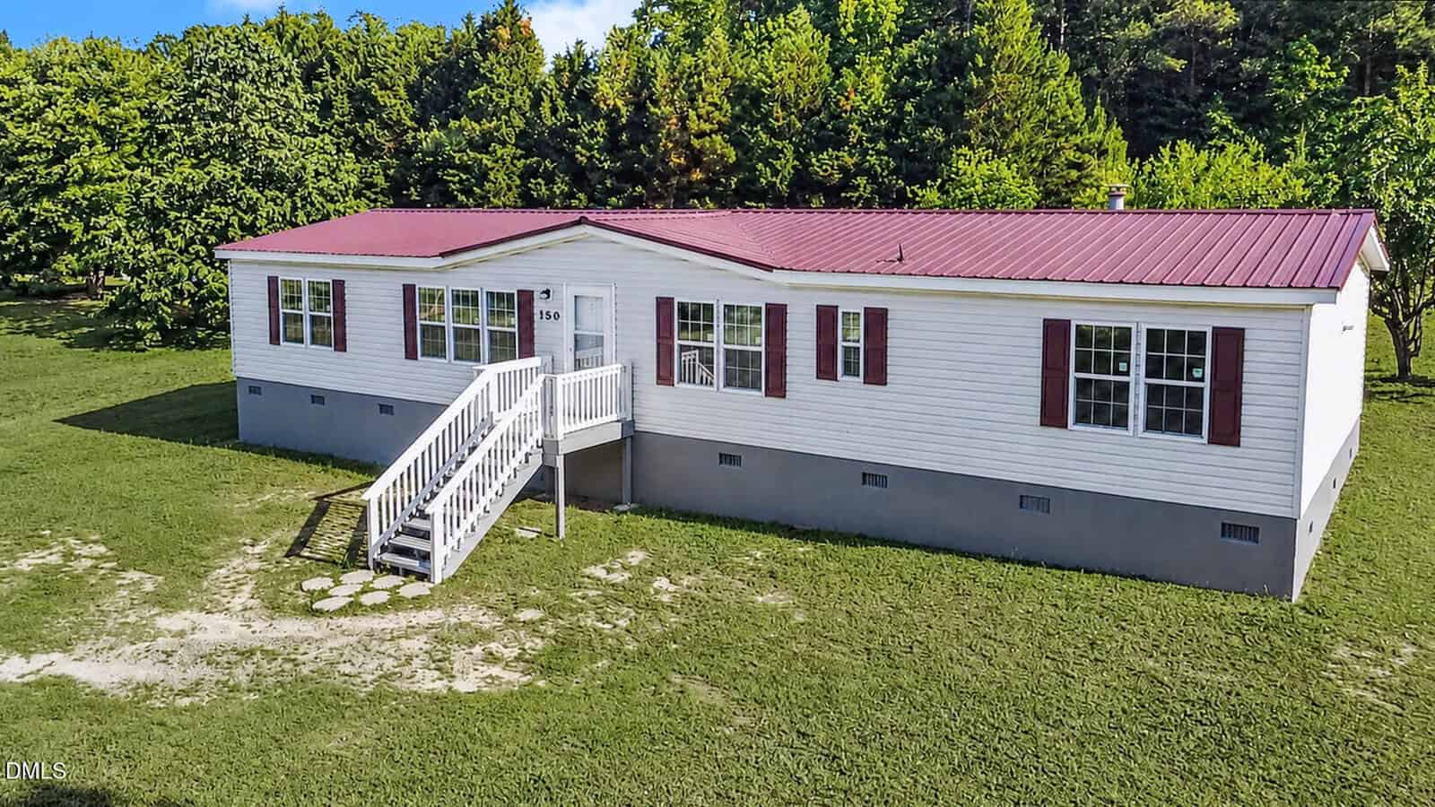 A single-story white manufactured home with a red metal roof and red shutters, surrounded by green grass and trees. A small staircase leads up to the front door.