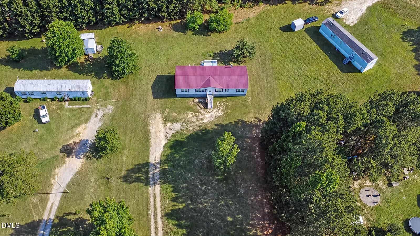 Aerial view of a rural property with three houses, one with a red roof at the center, surrounded by grass, trees, and a few outbuildings, with a dirt driveway curving through the area.