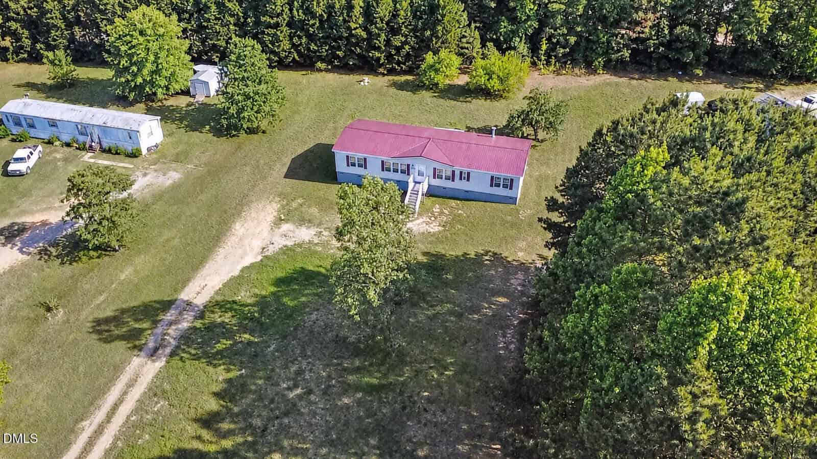 Aerial view of a red-roofed house and a white mobile home on a grassy lot with trees surrounding the property, a dirt driveway, and a small white shed in the background.
