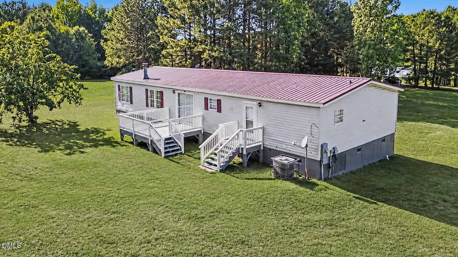 A white single-story manufactured home with a red metal roof sits on a large grassy lawn, surrounded by trees. The house has a front porch with steps and railings, and an air conditioning unit is visible on the side.