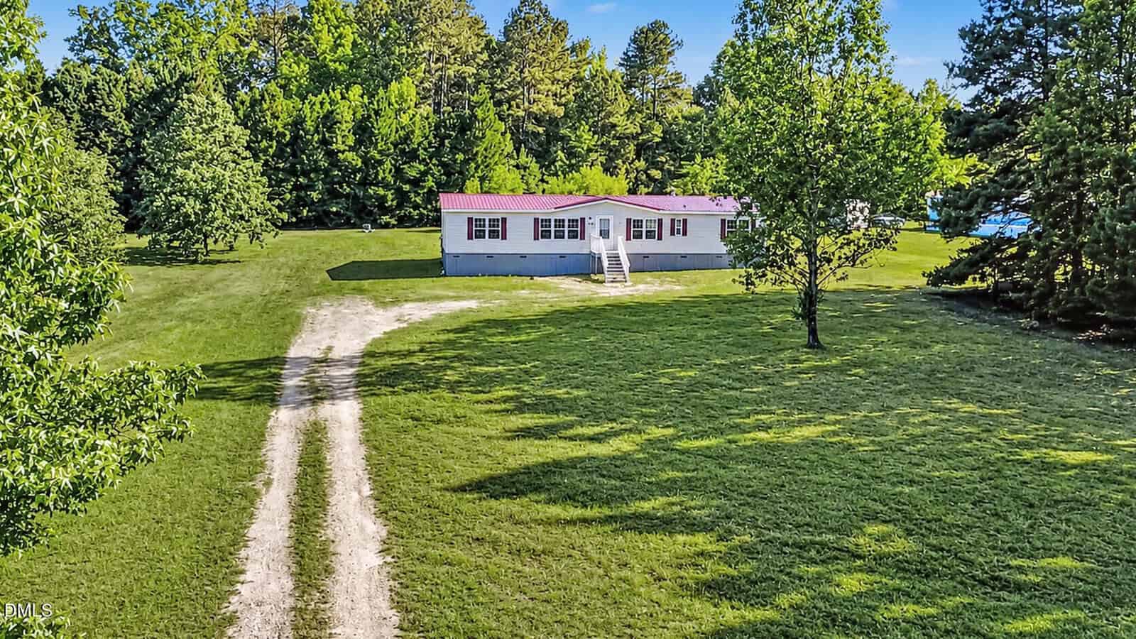 A single-story house with a red roof sits on a large, grassy lawn. A gravel driveway leads up to the front steps, and trees surround the property under a clear blue sky.