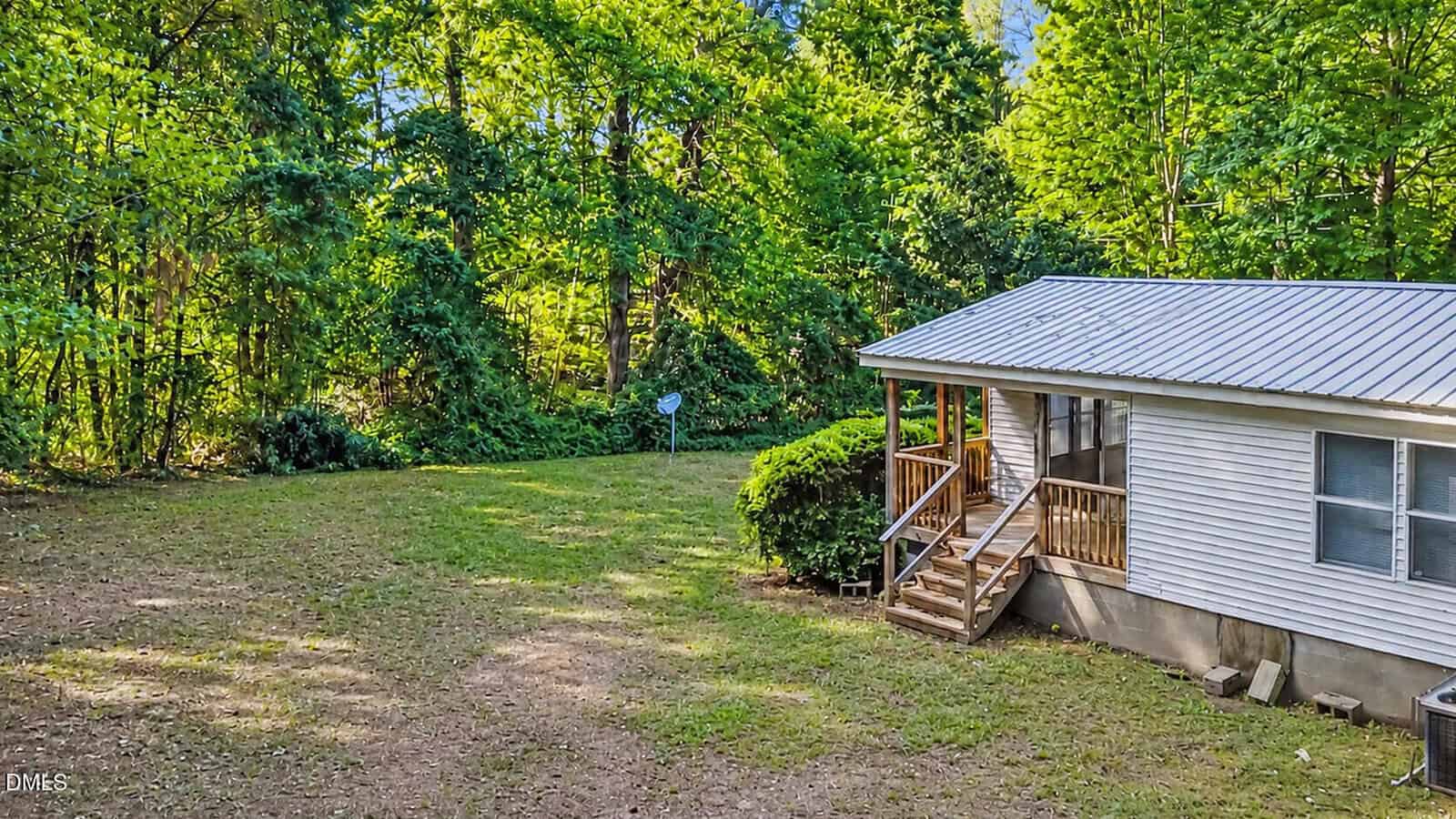 A small white house with a metal roof and a wooden porch sits beside a grassy yard, surrounded by dense green trees and shrubs under bright sunlight.