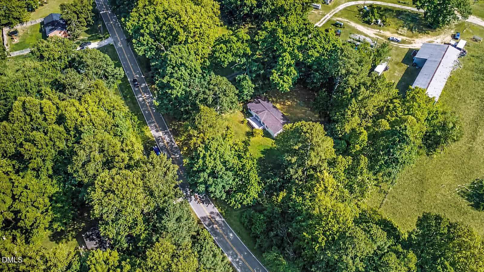 Aerial view of a rural area with a house surrounded by dense trees, a winding road, and several outbuildings nearby. Lush green vegetation covers most of the landscape.