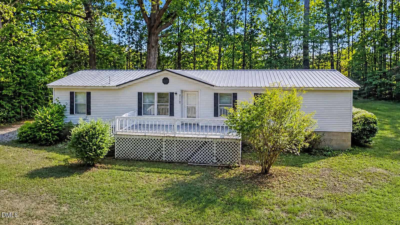 Charming single-story white house with a metal roof on 870 Jackson Royster Road in Henderson, featuring a front porch with white railing, steps to the yard, and surrounded by bushes and trees with a wooded backdrop.