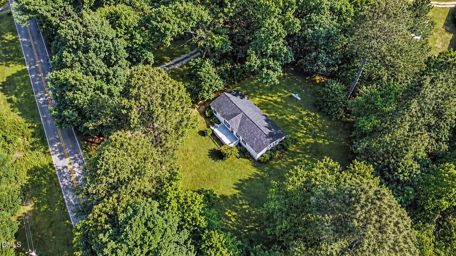 Aerial view of a small house with a gray roof surrounded by a large, grassy yard and dense green trees, next to a narrow road on the left side of the image.