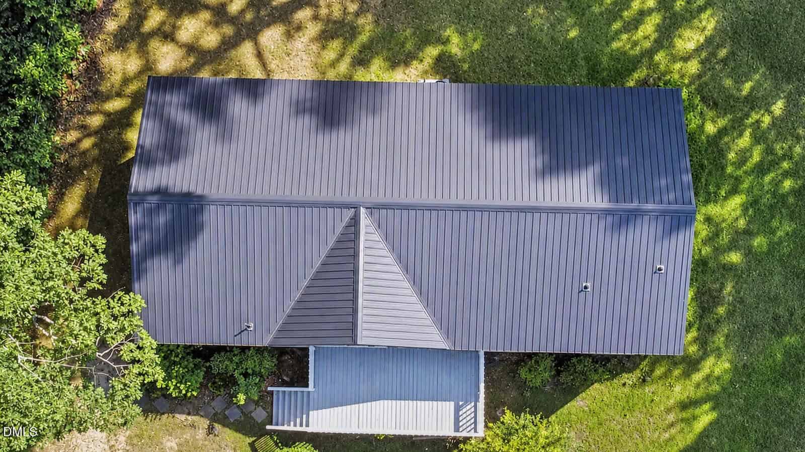 Aerial view of a house with a dark metal roof, surrounded by green grass and trees. Shadows from trees partially cover the roof, and a small covered porch is visible at the front.
