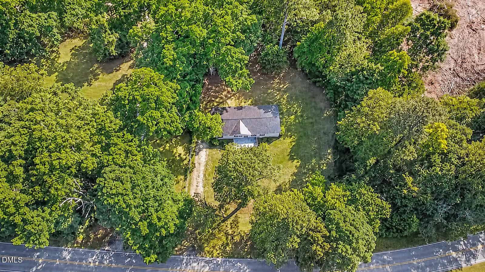 Aerial view of a small house surrounded by dense green trees and grass, with a driveway leading to a road at the bottom of the image.