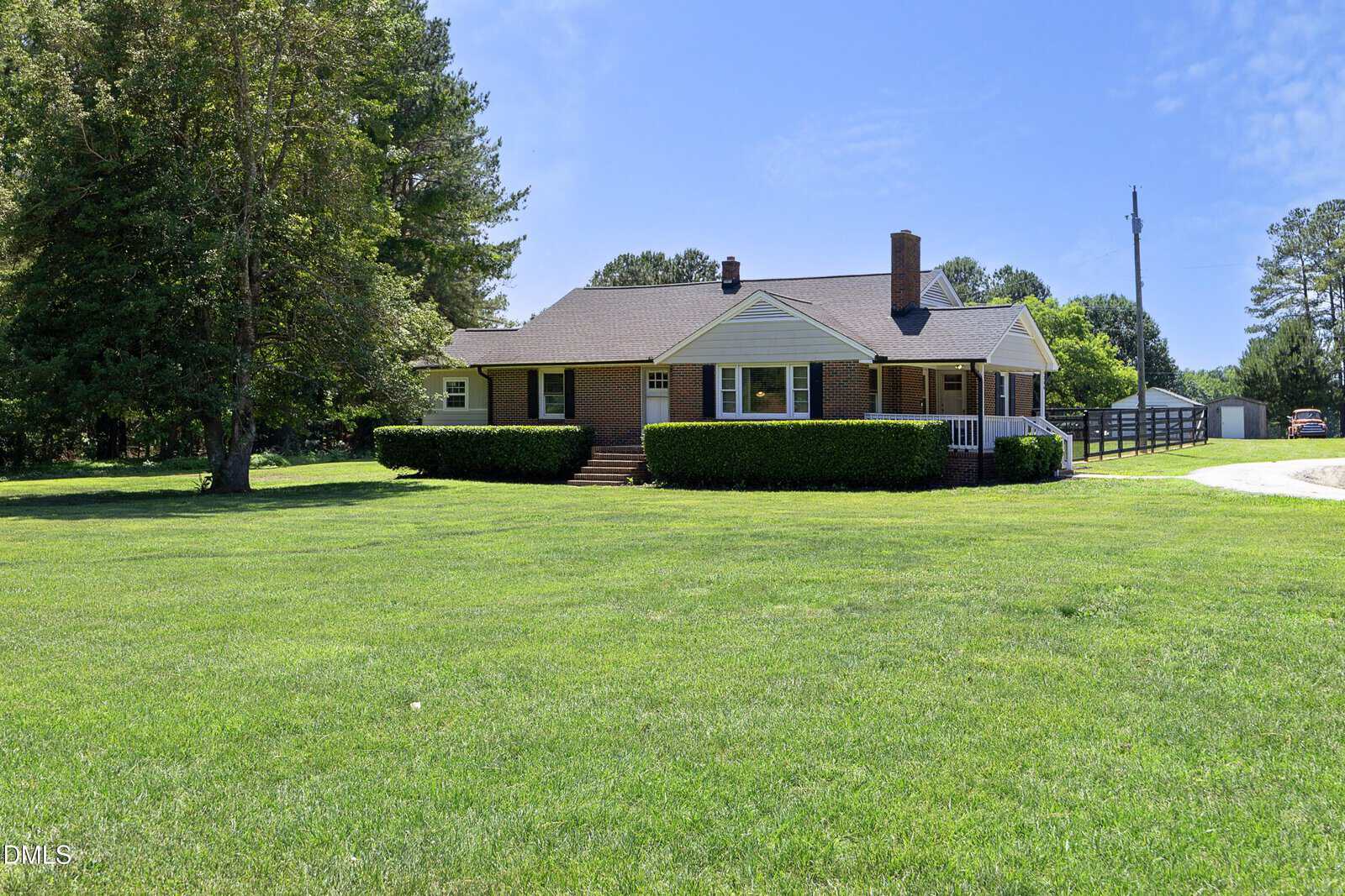 A single-story brick house with a porch and chimney sits on a large, grassy lawn, bordered by neatly trimmed hedges. Tall trees and a clear blue sky are in the background.