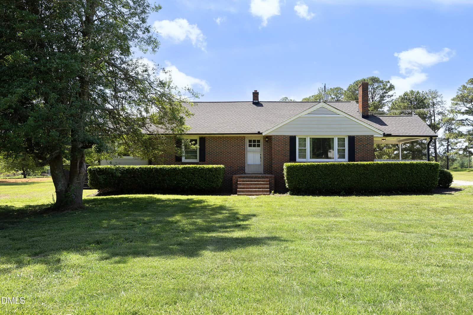 A one-story brick house with white trim and a gabled roof at 4306 Tommie Sneed Road, Oxford, features central front steps and manicured hedges on a large grassy lawn beneath a blue sky with scattered clouds.