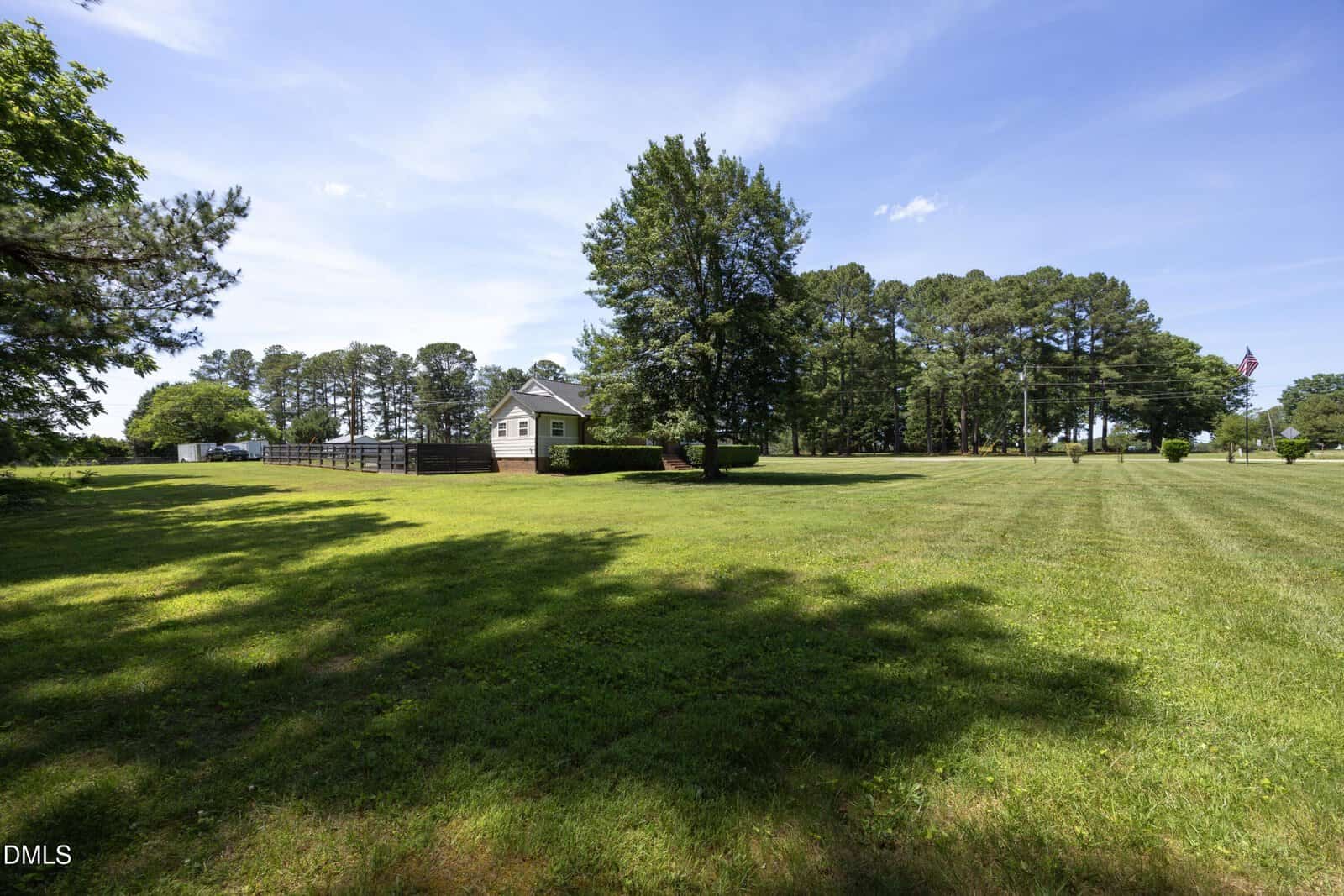 A small white house sits on a large, grassy lawn with tall trees in the background under a blue sky with a few clouds. The area appears peaceful and spacious.