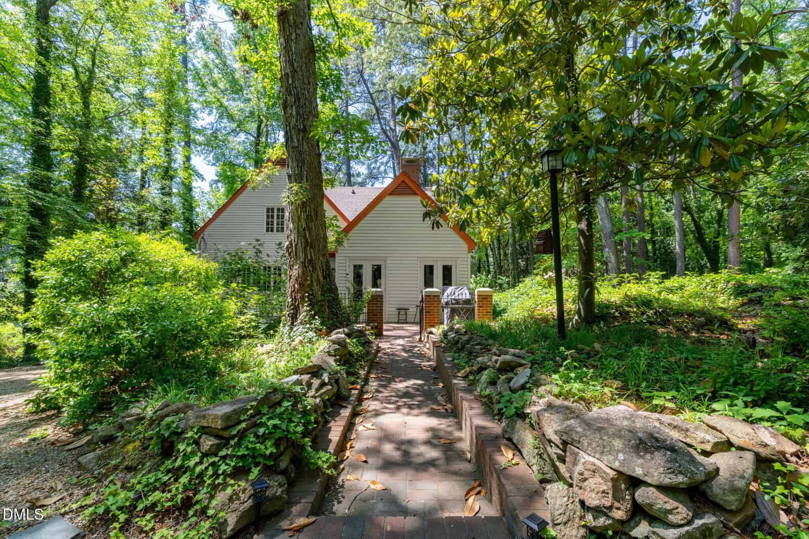 A cottage with white siding and an orange roof sits among lush green trees and plants. A brick path lined with stone walls leads to the front door. Sunlight filters through the leaves, creating a serene, forested setting.