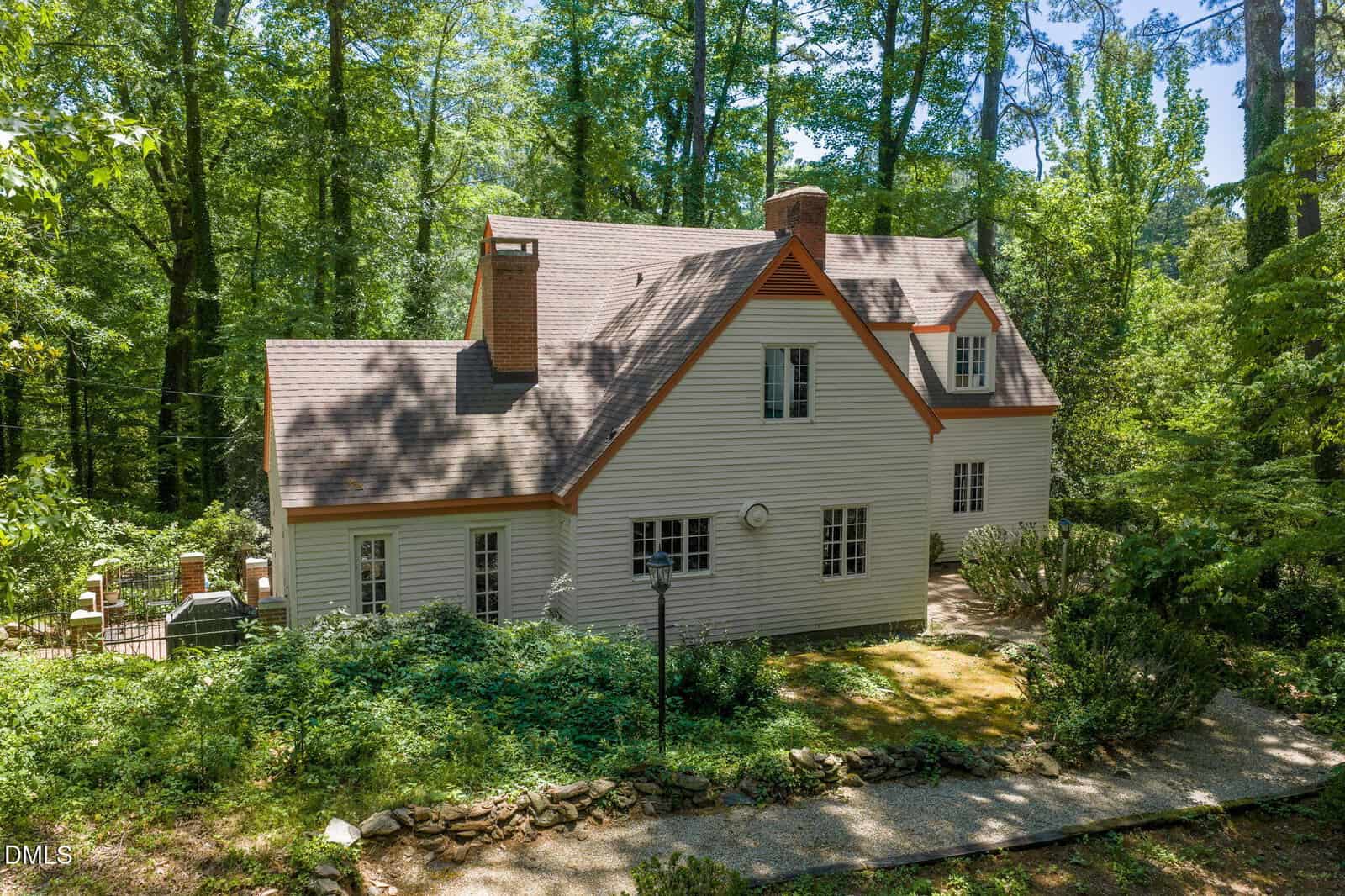 Two-story white house with multiple chimneys and gabled roof, surrounded by dense green trees and lush landscaping on a sunny day. A stone path leads to the entrance.