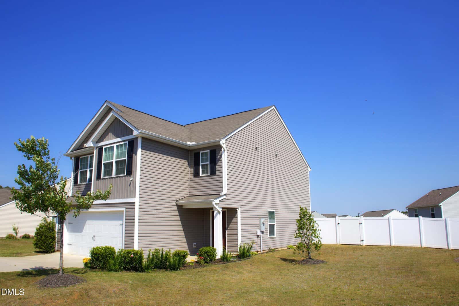 A two-story beige suburban house with white trim, a double garage, and a small covered entrance, surrounded by a white fence and minimal landscaping, under a clear blue sky.