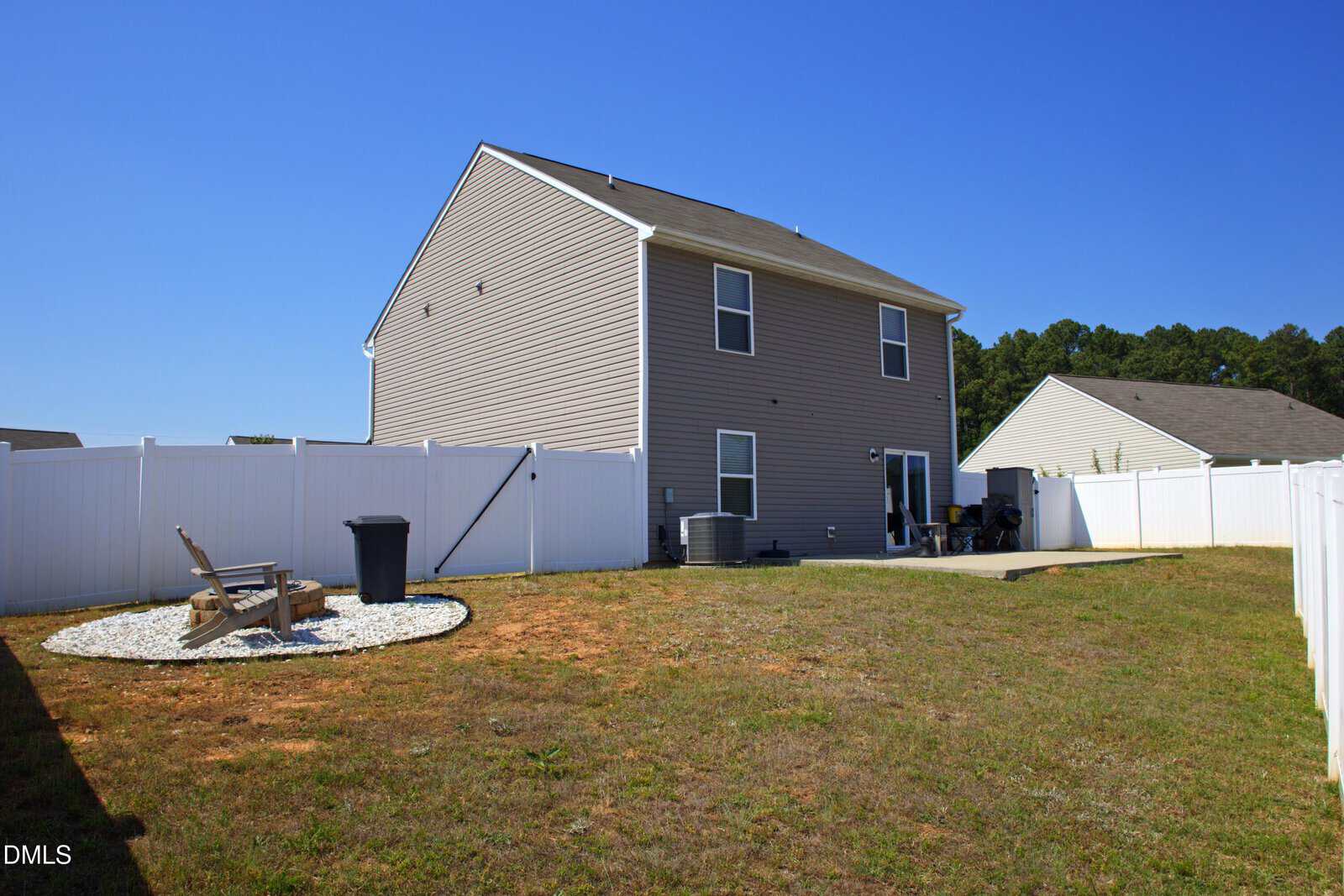 A fenced backyard with a two-story beige house, patio, air conditioning units, and a small fire pit area with chairs and white gravel. The grass is patchy, and the sky is clear and blue.