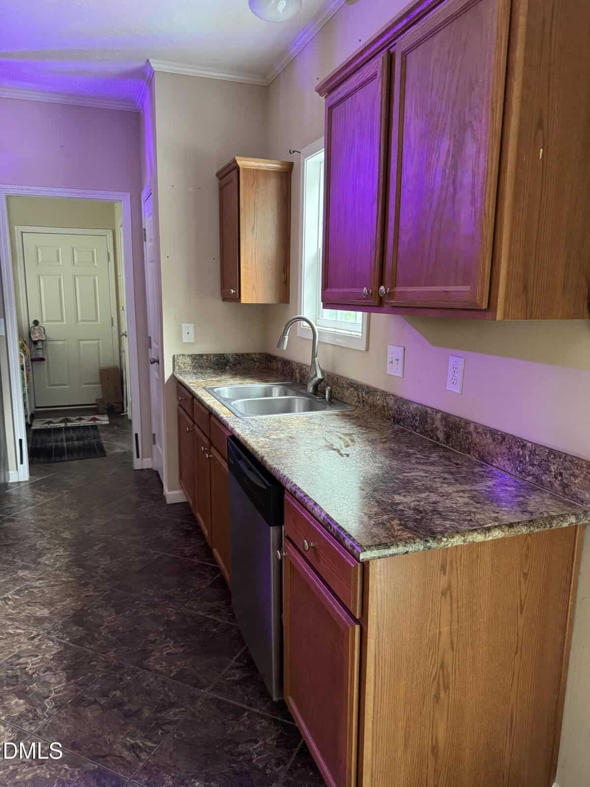 Narrow kitchen with brown cabinets, marble-patterned countertops, a double sink, dishwasher, and tile floor. A window above the sink lets in light. Purple light is visible on the ceiling and cabinets. Doorway at the far end.