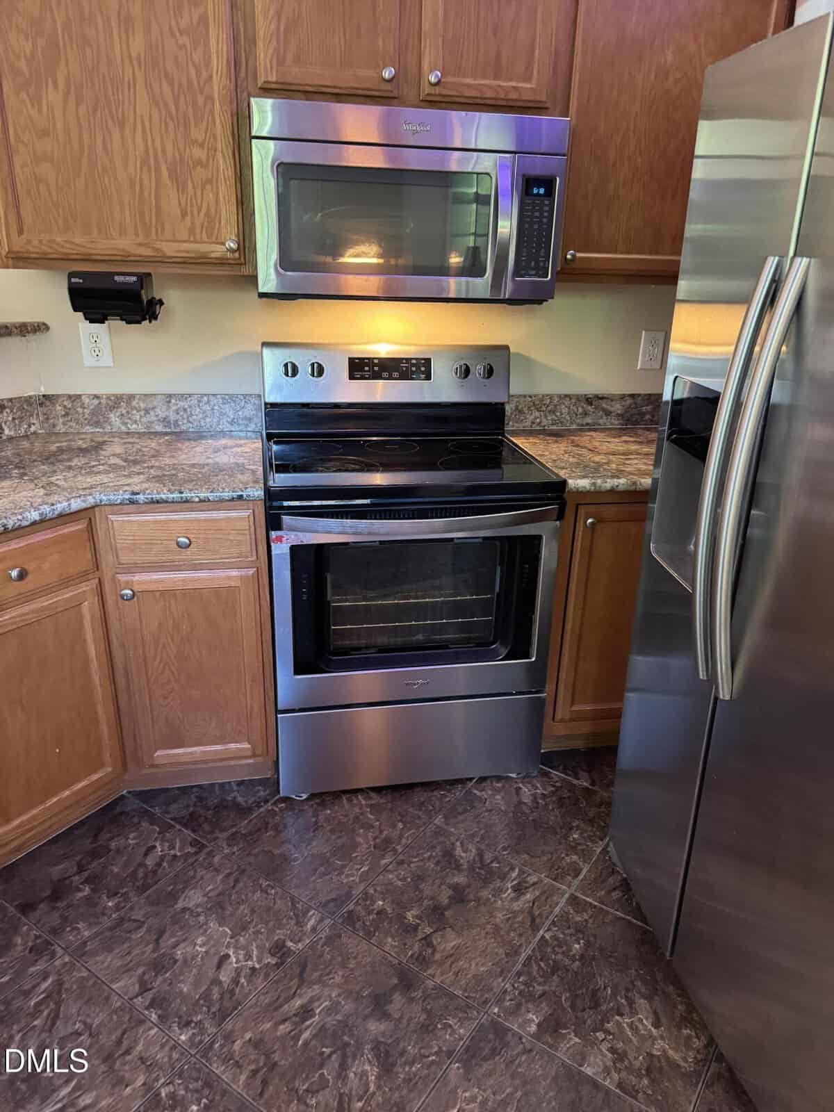 A stainless steel oven with a glass door is below a built-in microwave in a kitchen with brown cabinets, granite-patterned countertops, and a stainless steel refrigerator to the right.
