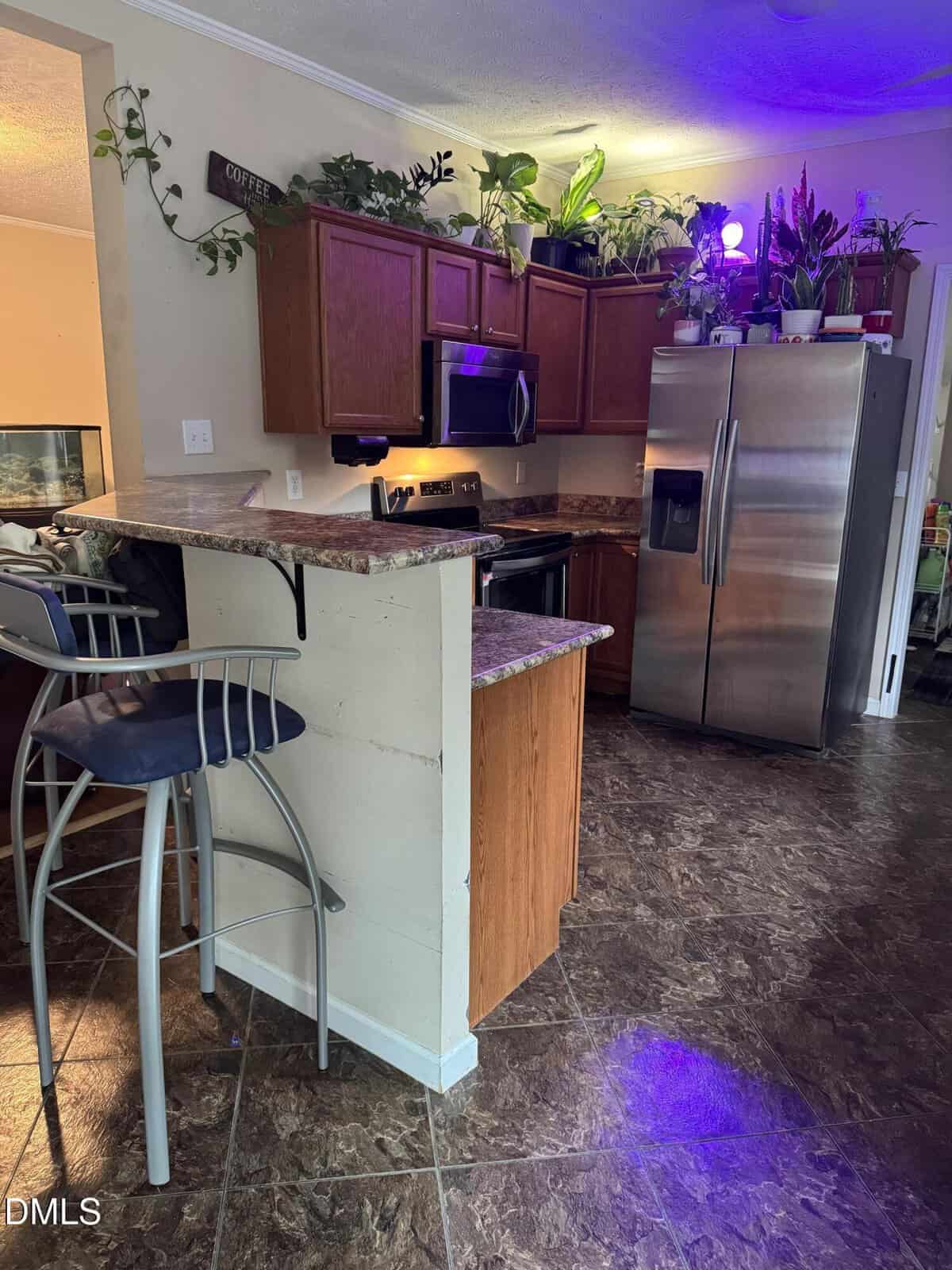 A kitchen with brown cabinets, stainless steel appliances, a breakfast bar with two stools, and several potted plants on top of the cabinets. The floor is tiled, and purple grow lights are illuminating some plants.