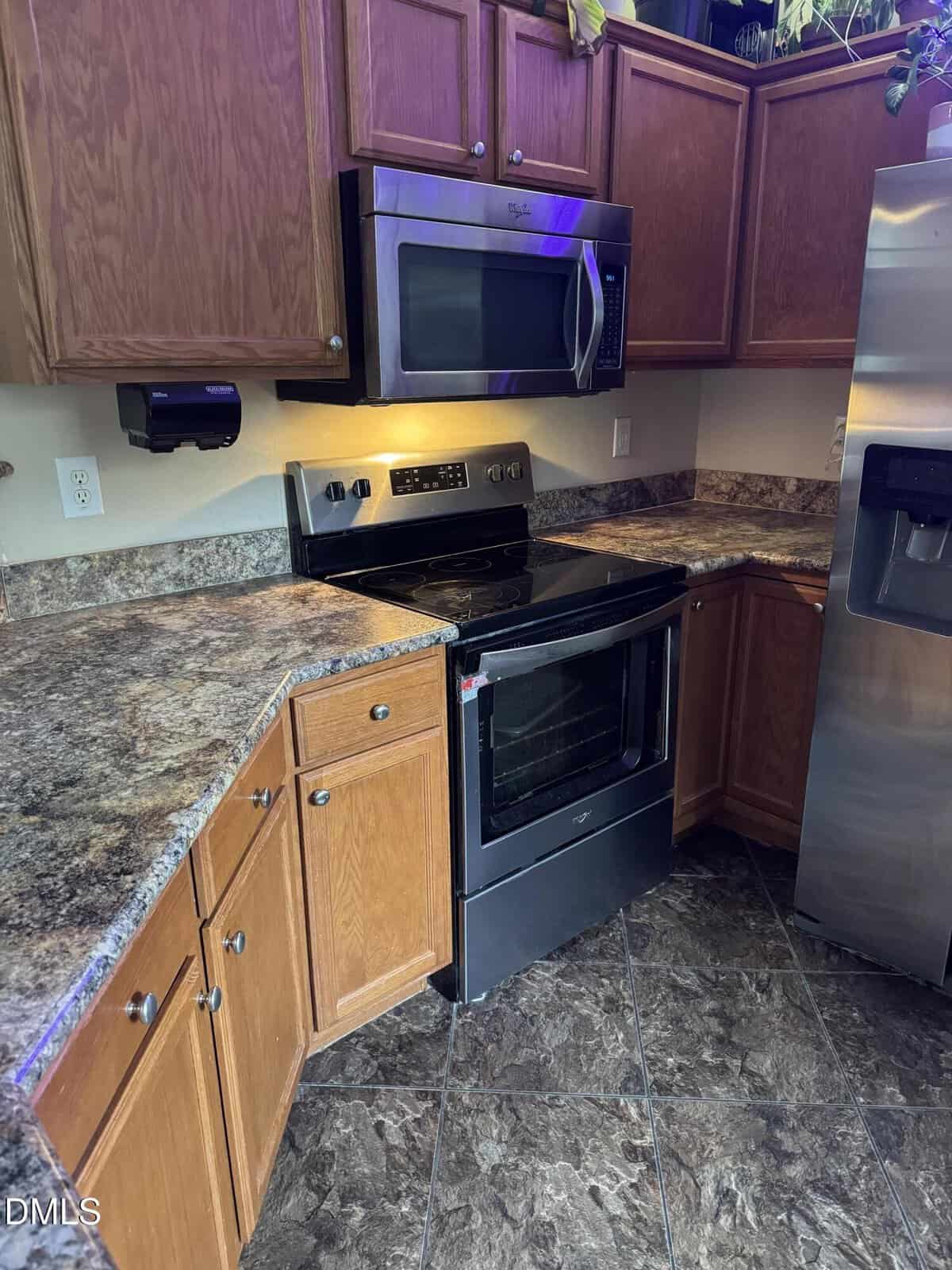 Modern kitchen with brown wood cabinets, marble-pattern countertops, stainless steel oven, microwave, and fridge, and dark tile flooring. Small plants are on the upper cabinets.