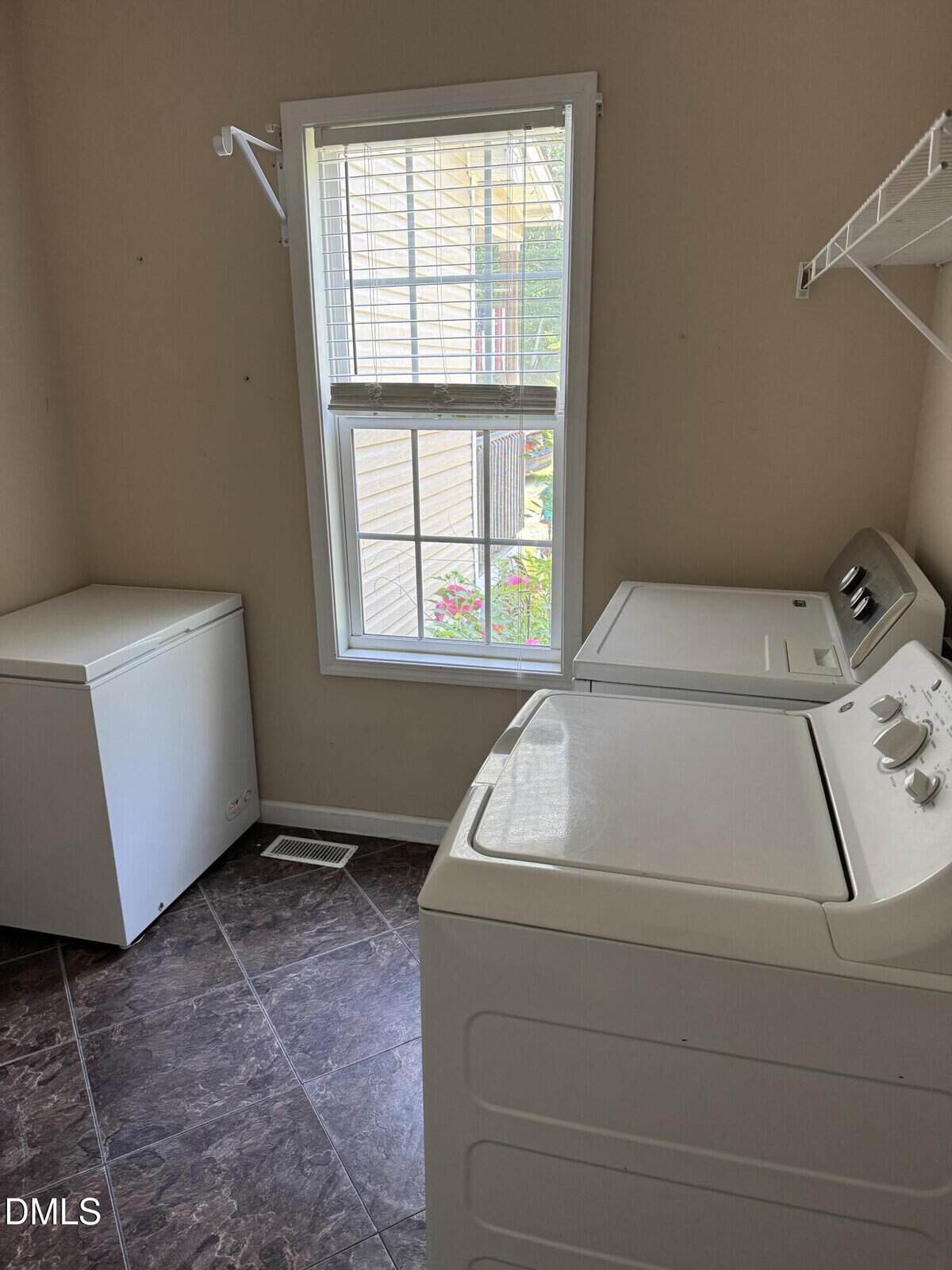 A laundry room with a chest freezer, a washing machine, a dryer, a window with blinds, and a wire shelf on the wall.