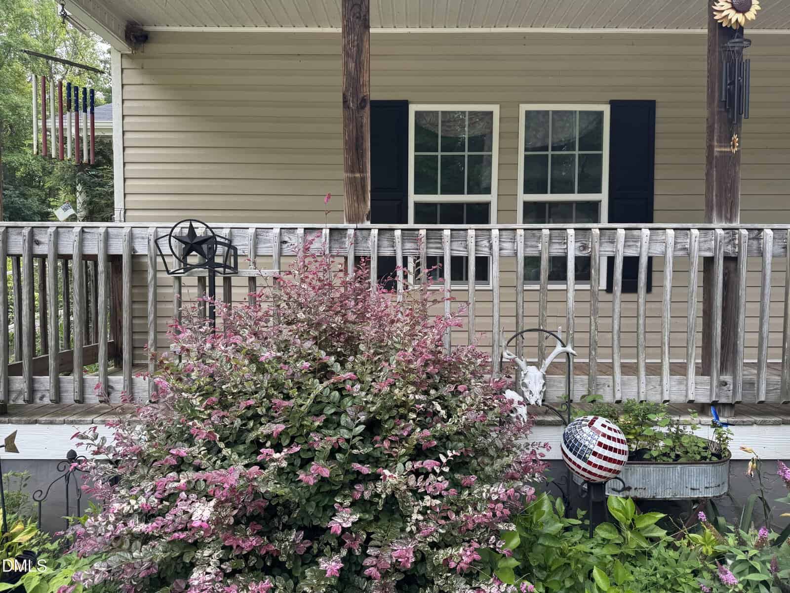 A bush with pink and green leaves grows in front of a wooden porch with two windows. The porch has a weathered railing, patriotic decorations, and a hanging wind chime. The house exterior is tan with black shutters.