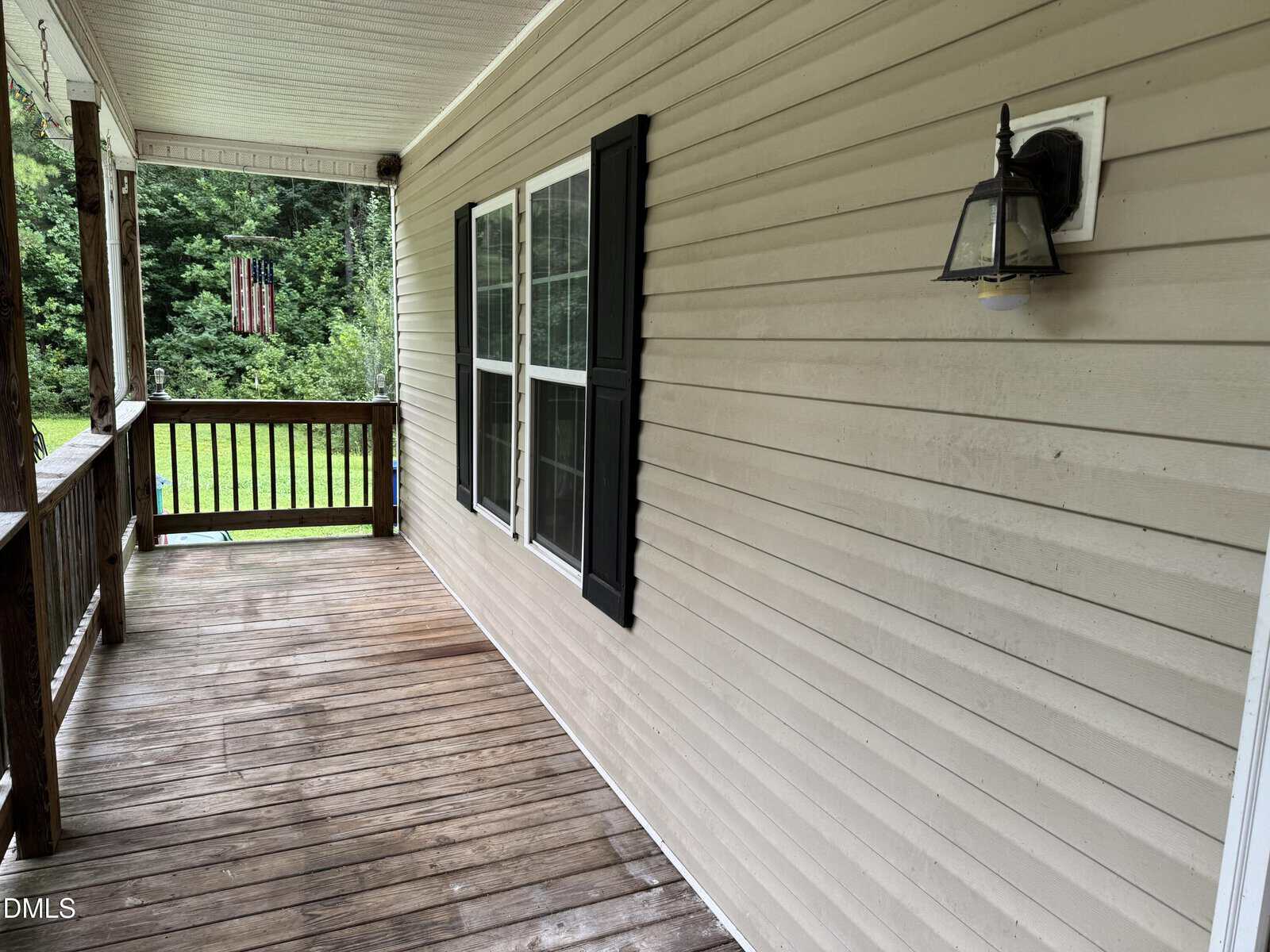 A wooden porch with a tan siding wall, black shutters on the windows, and a lantern-style light fixture. Trees and an American flag are visible in the background.