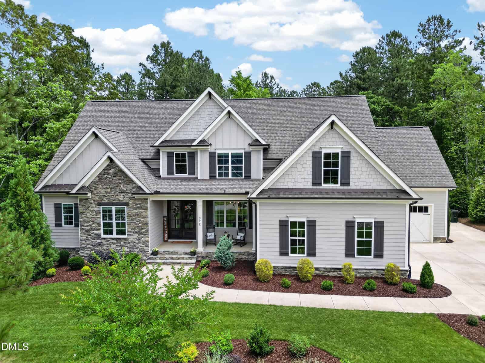 A two-story modern house at 3626 River Watch Lane, Franklinton, features gray siding, stone accents, black shutters, and a covered porch. Lush landscaping and trees surround the home, with a driveway leading to a side-entry garage.