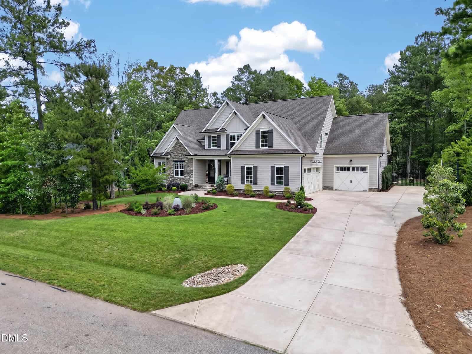 Large two-story house with gray siding, white trim, gabled roof, and attached garage. Well-manicured lawn, curved driveway, and landscaped garden beds surrounded by tall trees and blue sky.