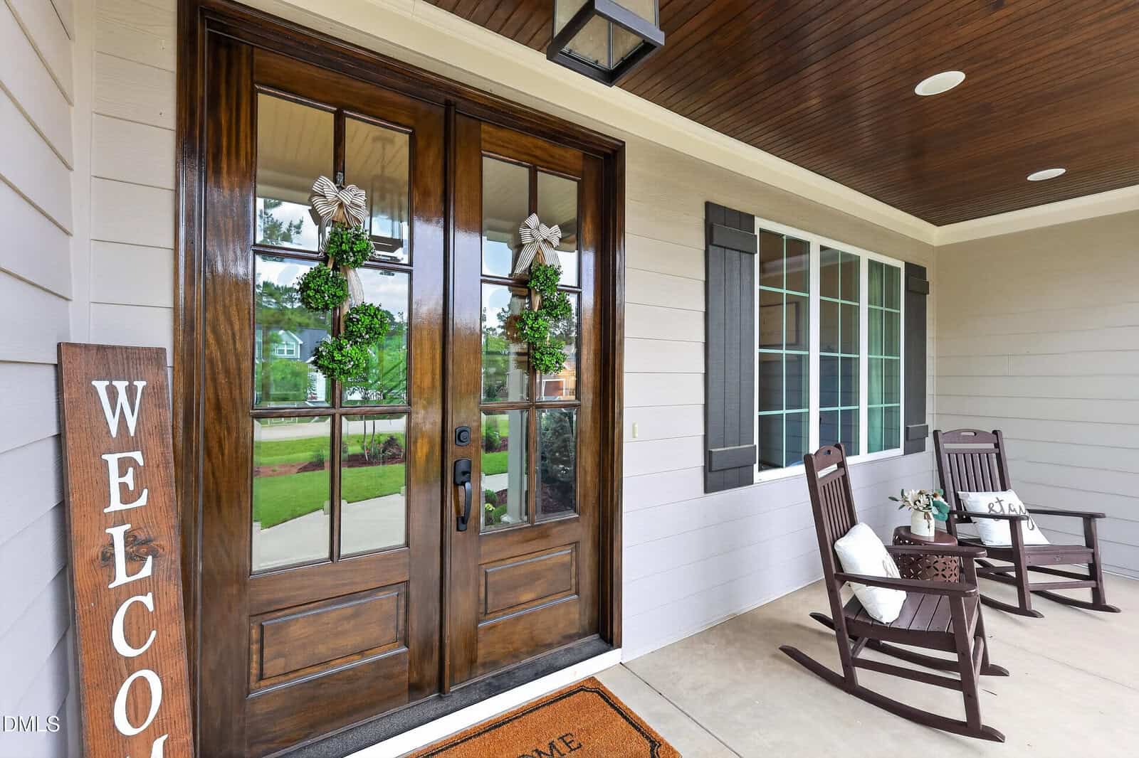 A front porch with double wooden doors decorated with wreaths, a "Welcome" sign, two rocking chairs with cushions, a small table with mugs, and large windows under a wood-paneled ceiling.
