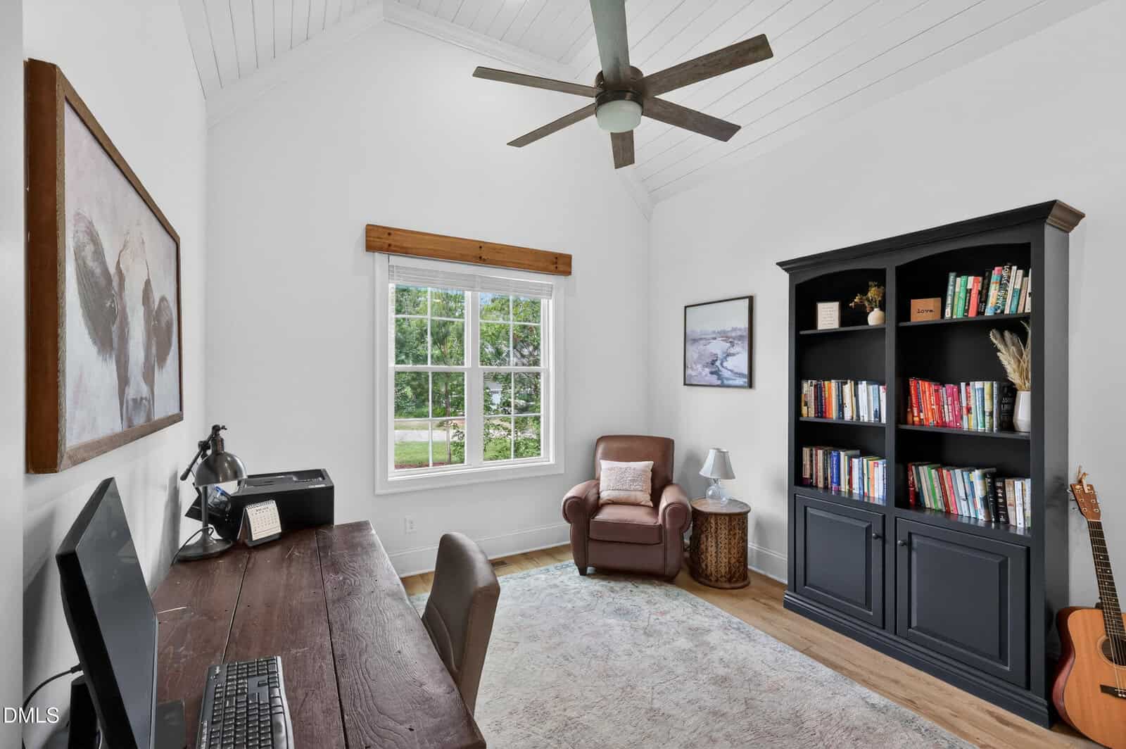 Home office with a wooden desk, computer, and chair on the left, armchair by a window in the center, black bookshelf on the right, guitar by the bookshelf, art on the walls, ceiling fan, and white walls with wood floors.