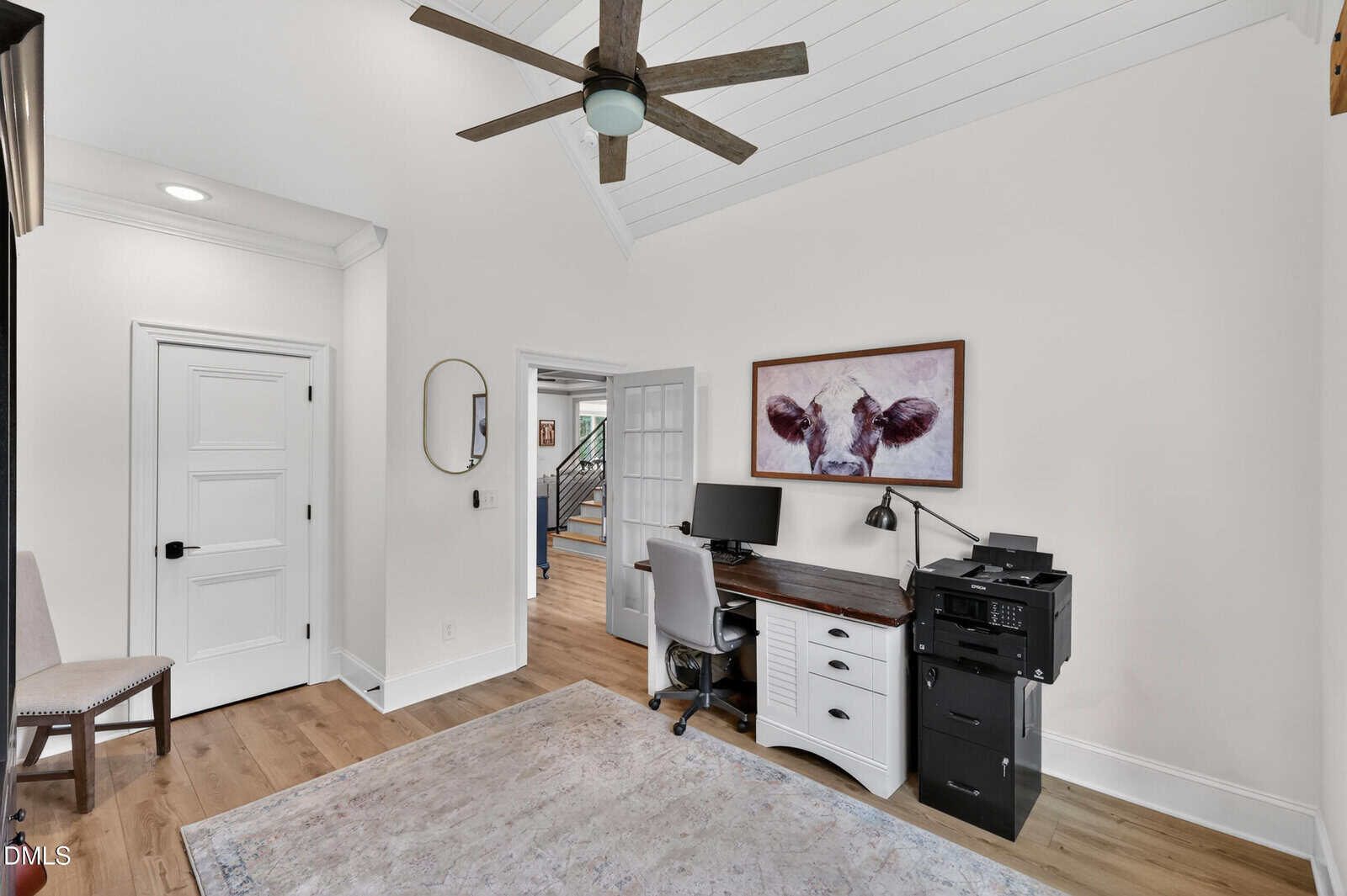 A home office with white walls, wood floors, a ceiling fan, a desk with a computer and printer, a chair, and a framed cow picture above. A doorway leads to a hallway with stairs.